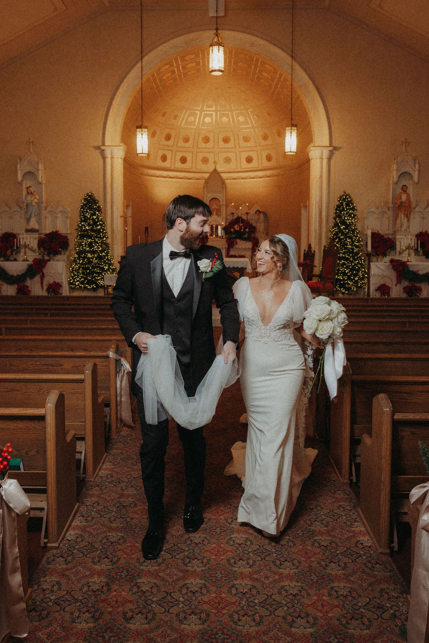 Bride and groom walk down church aisle together after ceremony past Christmas trees and wooden pews.