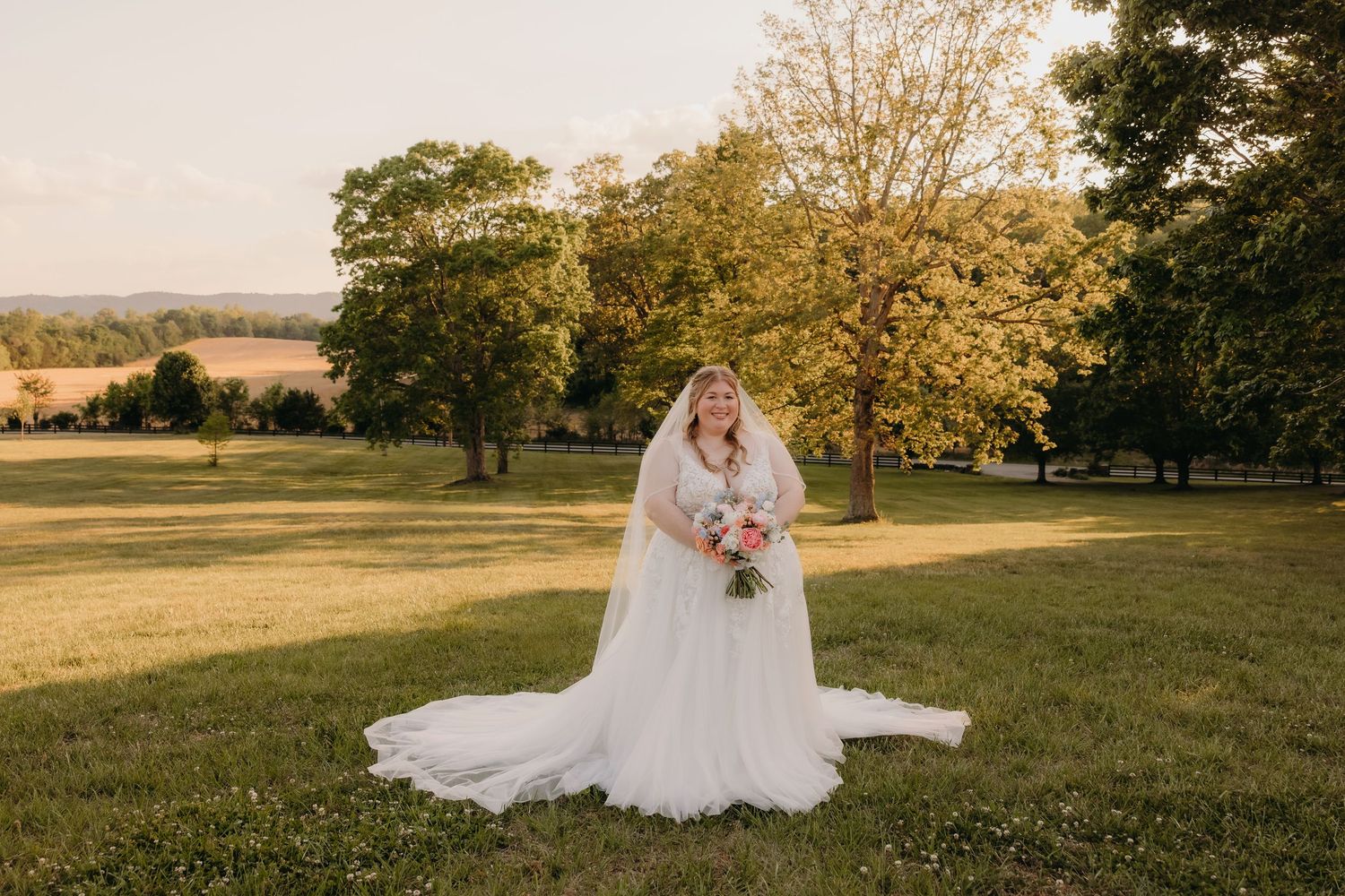 Bride in a flowing white gown and veil holds a bouquet while standing in a sunlit green field at golden hour.