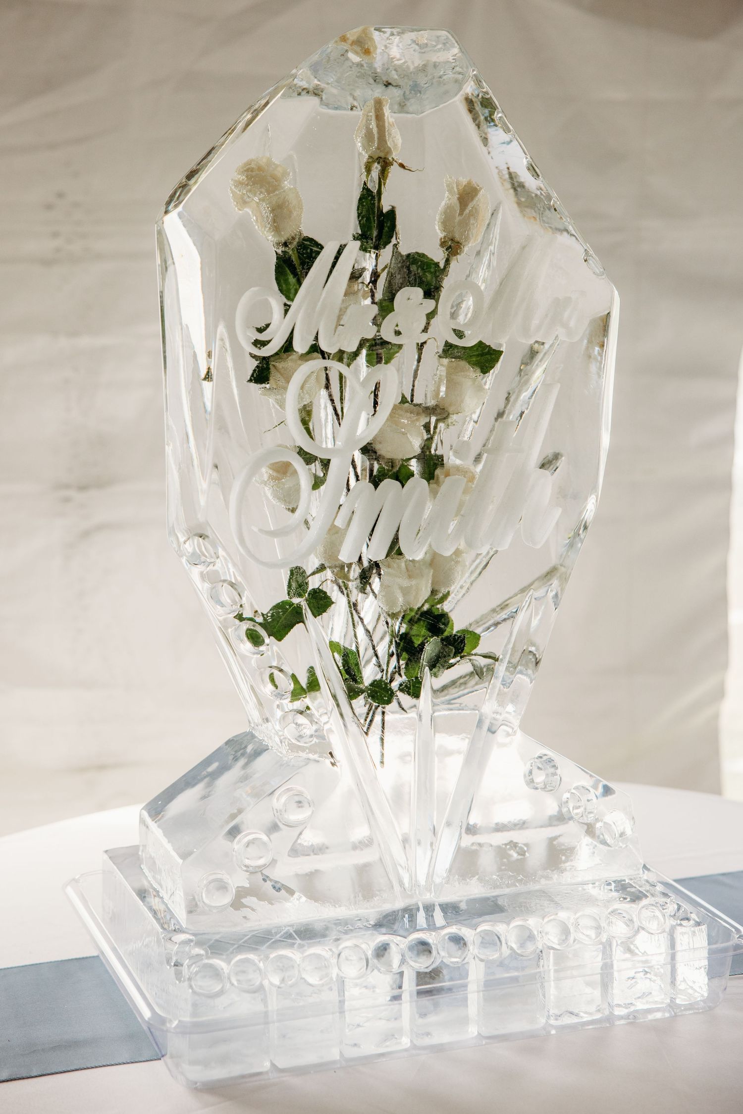 Elegant ice sculpture centerpiece engraved with wedding text and white roses at a wedding reception.