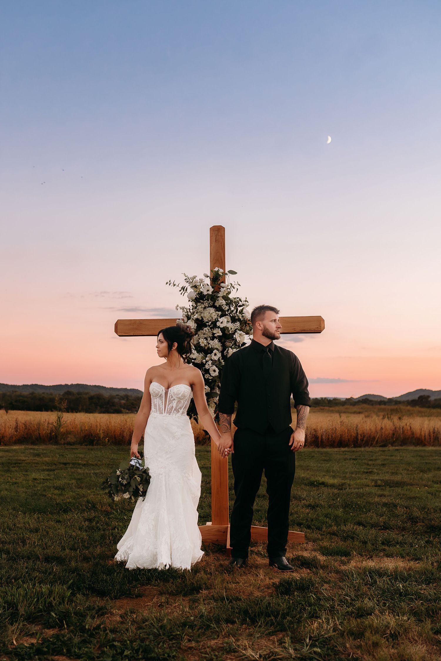 Bride and groom hold hands before a floral wooden cross at sunset during an outdoor wedding ceremony.