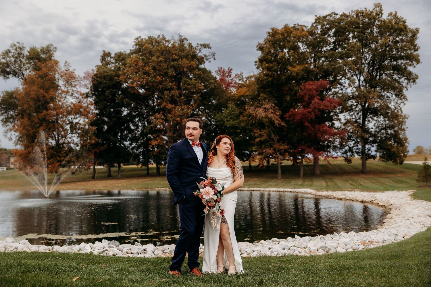 Bride and groom pose romantically in front of scenic pond surrounded by colorful fall foliage trees.