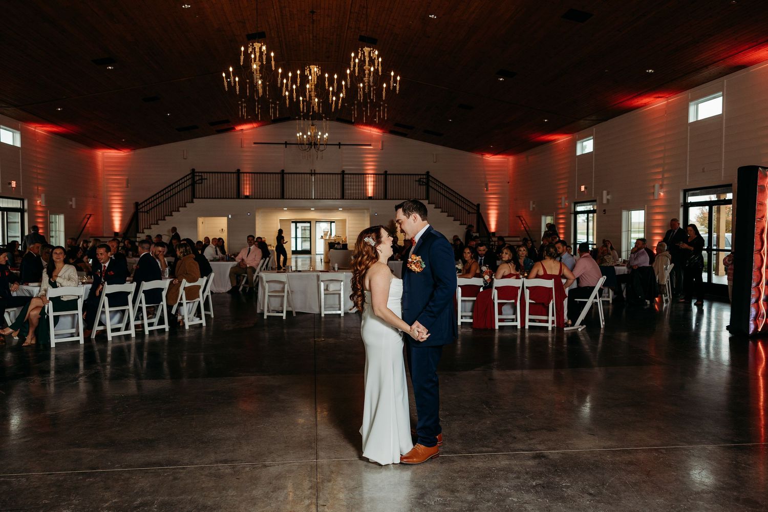 Bride and groom share first dance in elegant wedding venue with warm red uplighting and guests seated at decorated tables.