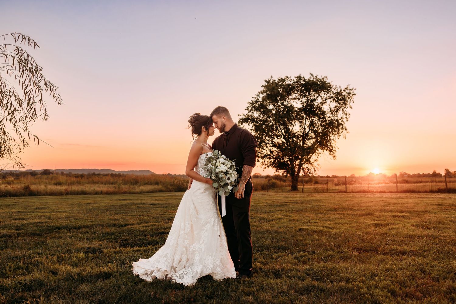 Bride and groom share a romantic sunset moment in a lush green field, with a glowing orange sky and tree backdrop.