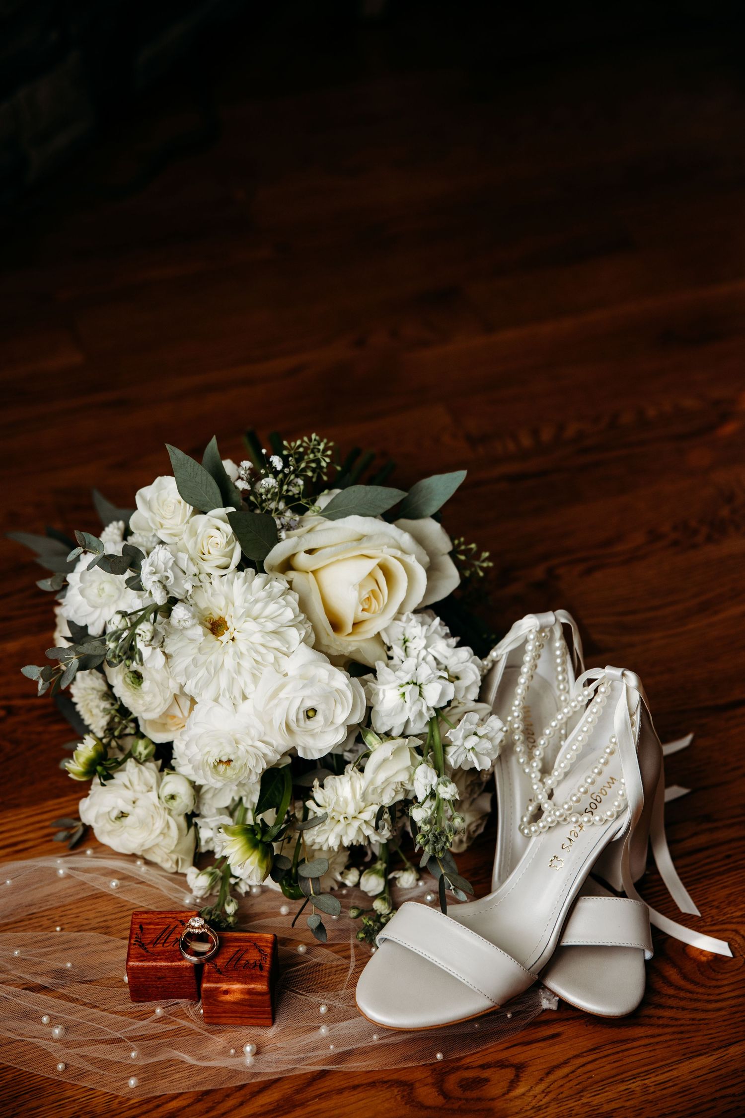 White bridal bouquet with roses beside elegant heeled wedding shoes and ring box on wooden floor.