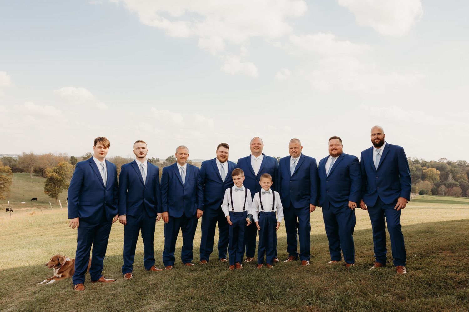 Groomsmen and groom posing outdoors in matching navy blue suits with a young ring bearer in suspenders on a wedding day.