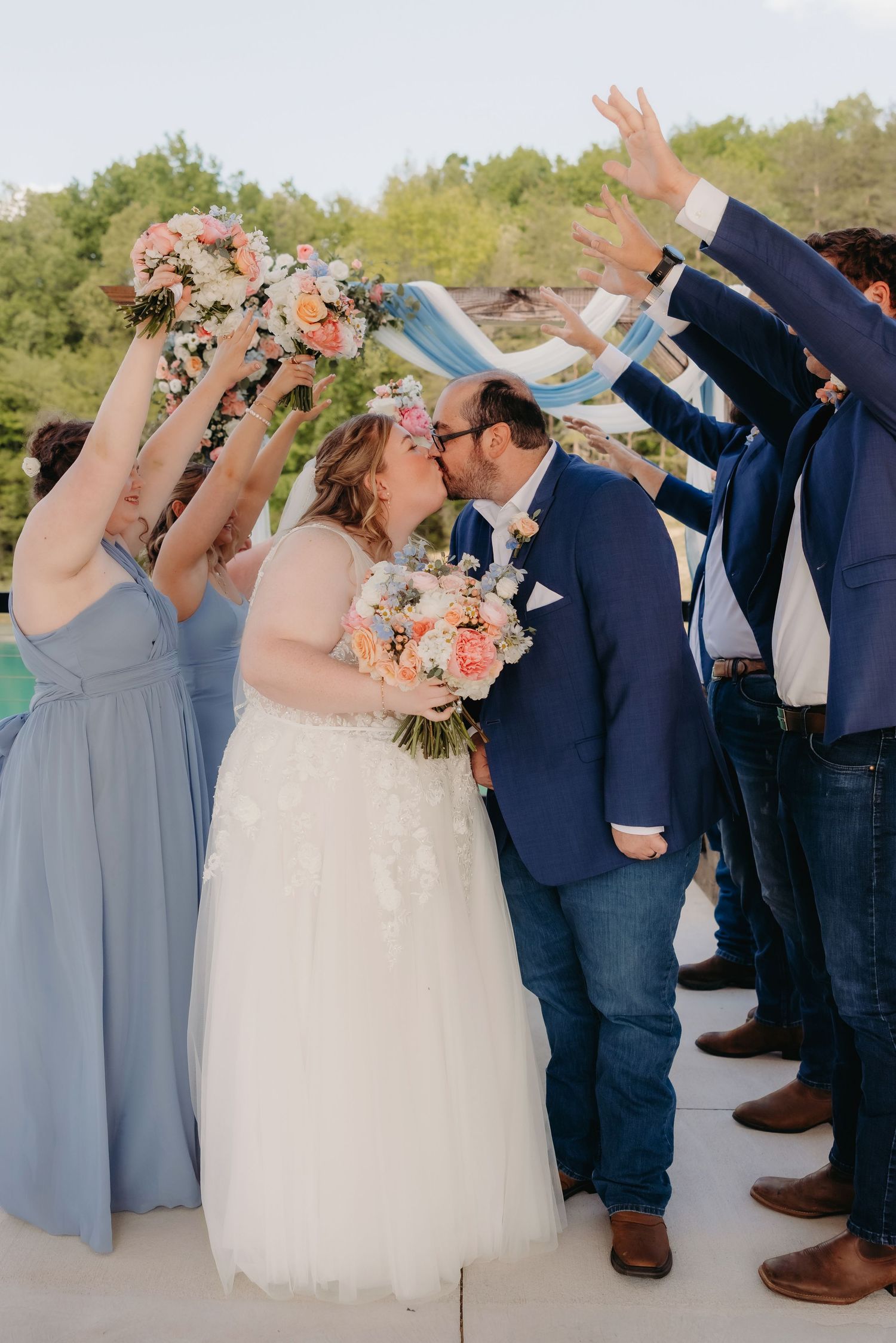 Bride and groom share a kiss while wedding party raises bouquets and ribbons in celebration outdoors.