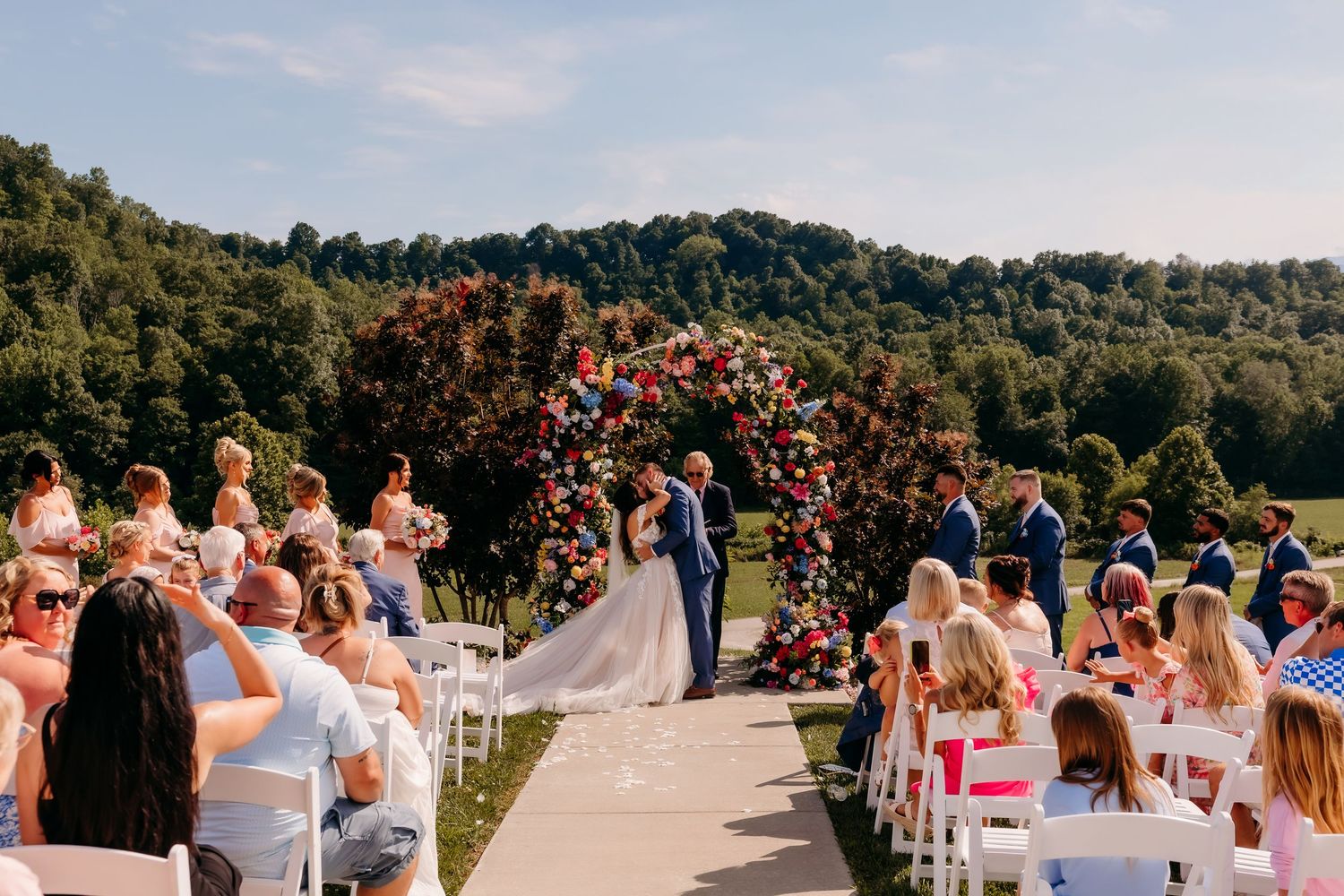 Outdoor wedding ceremony with floral arch, kissing couple, and scenic rolling hills backdrop.