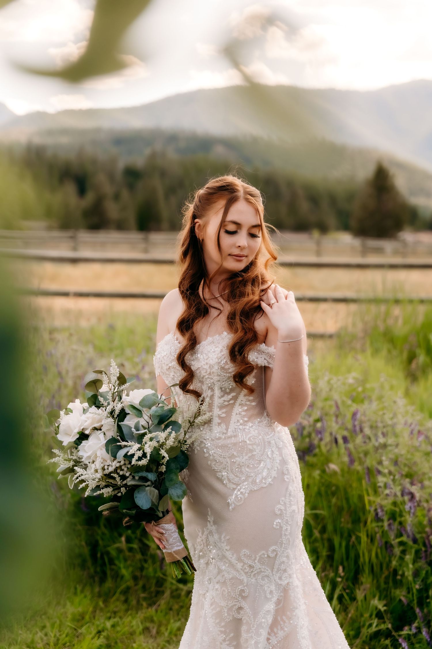 Bride in lace mermaid gown holds white bouquet in mountain meadow at golden hour.