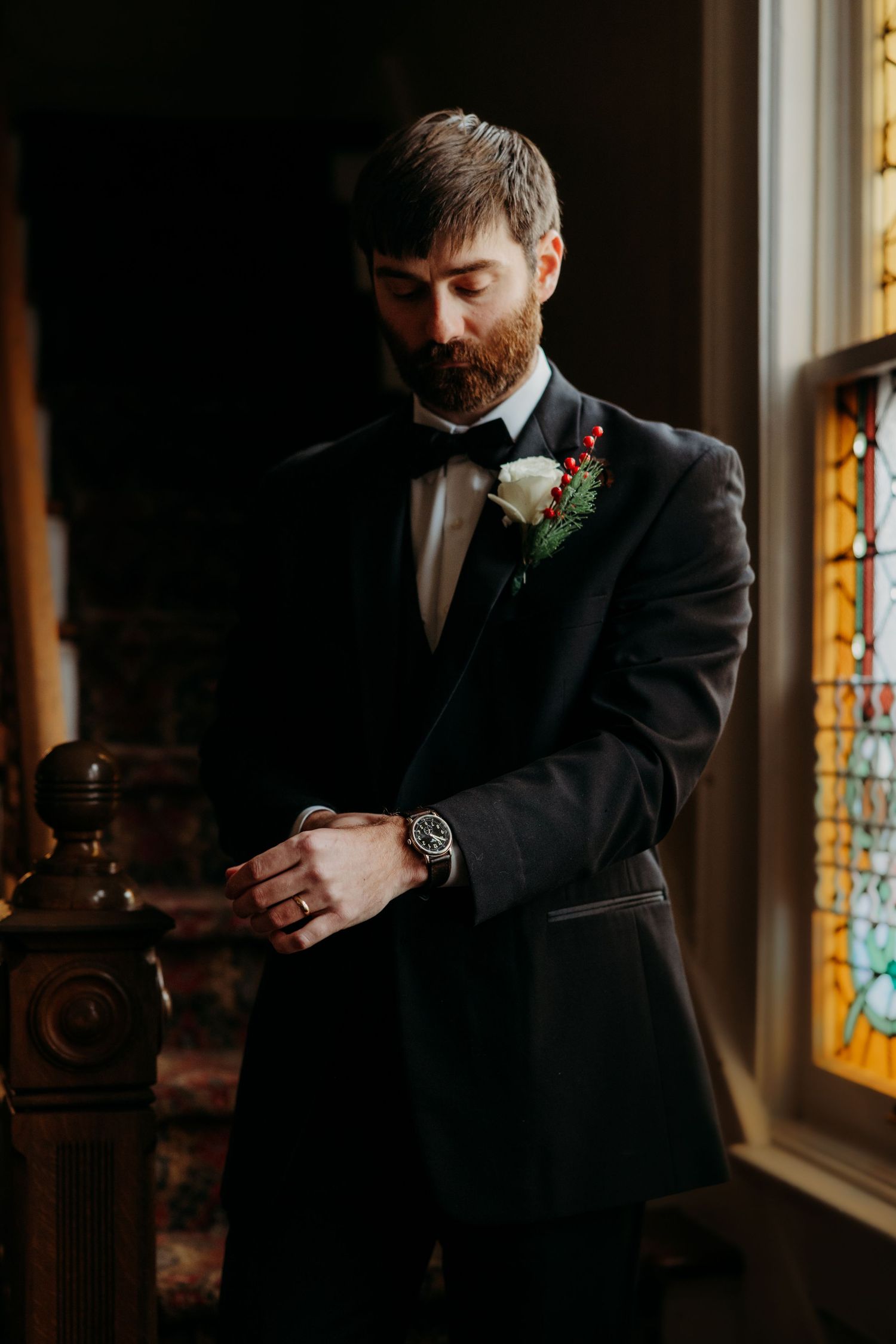 Dapper bearded groom in black tuxedo adjusts cufflinks beside stained glass window in moody lighting.