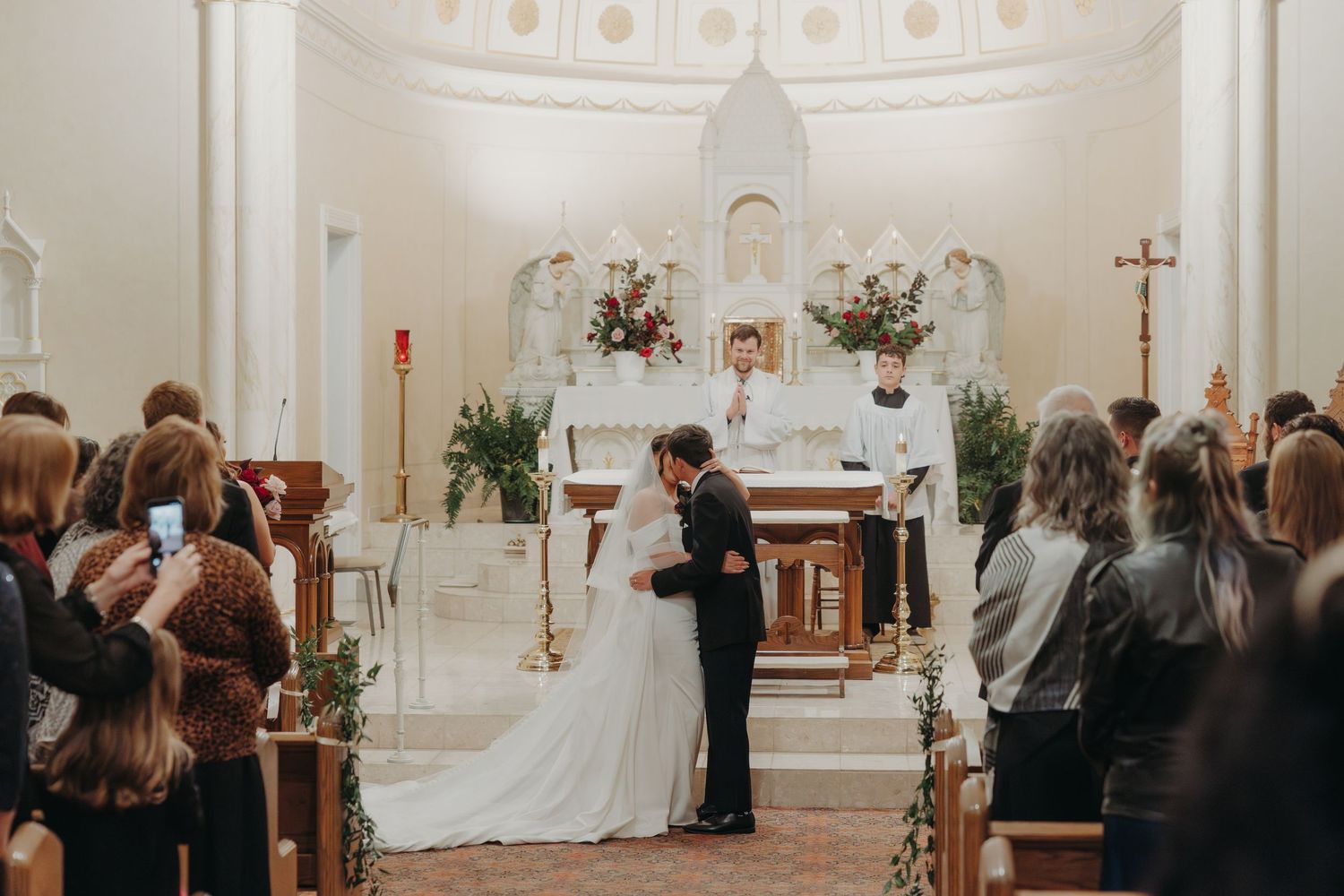 Bride and groom sharing a kiss at the altar during Catholic church wedding ceremony with guests watching and white floral decor.