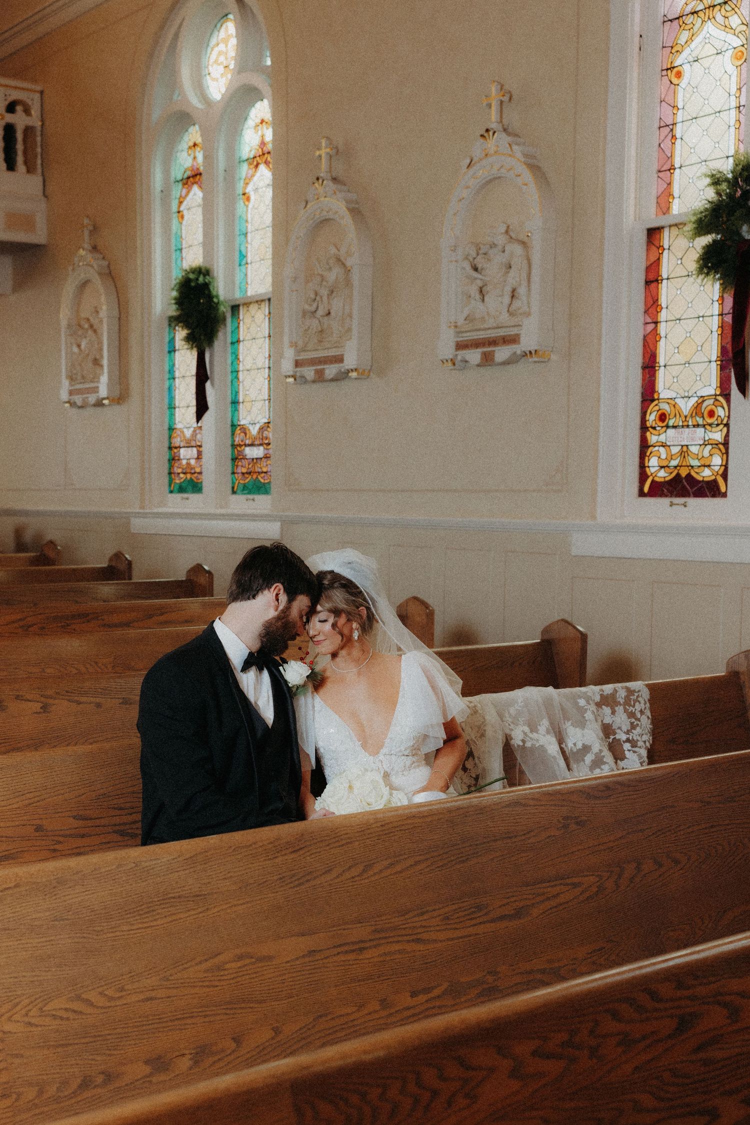 Bride and groom share intimate moment sitting together in wooden church pew beneath stained glass windows.