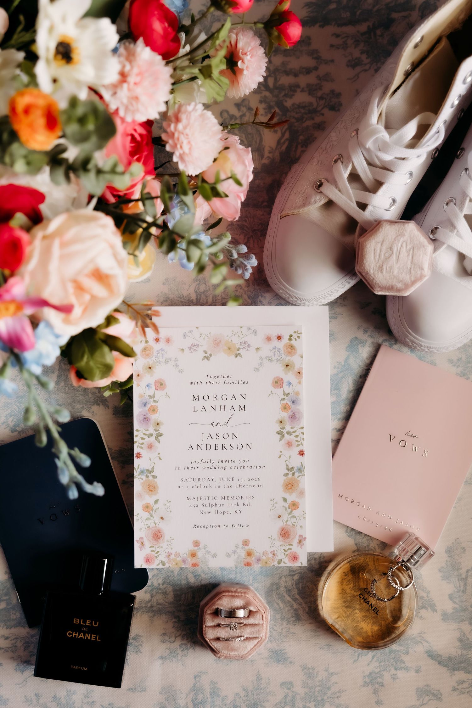 Wedding flat lay featuring floral invitation, rings, perfume, sneakers, and pink flowers on a decorative surface.