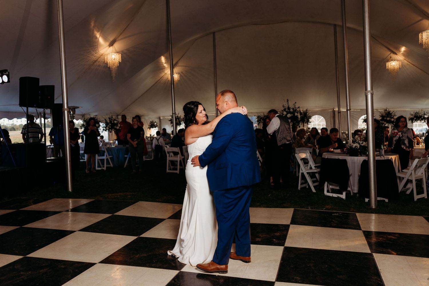 Bride and groom share their first dance on a black and white checkered floor under a wedding tent.