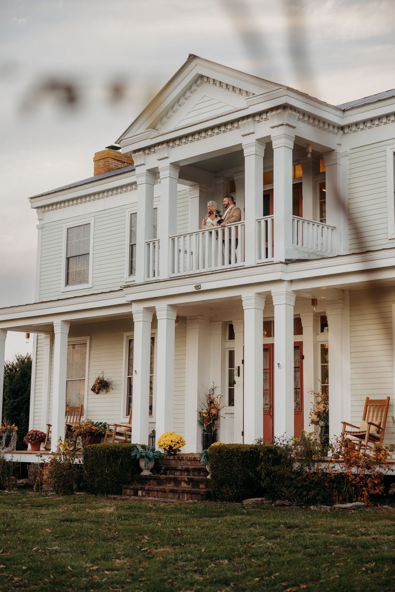 Elegant white Southern plantation-style wedding venue with couple on upper balcony at dusk.