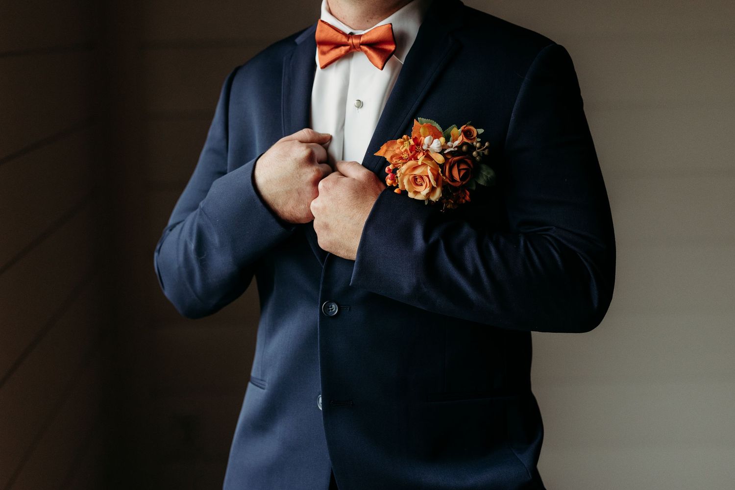 Groom in navy suit adjusting boutonniere with orange roses and matching bow tie.