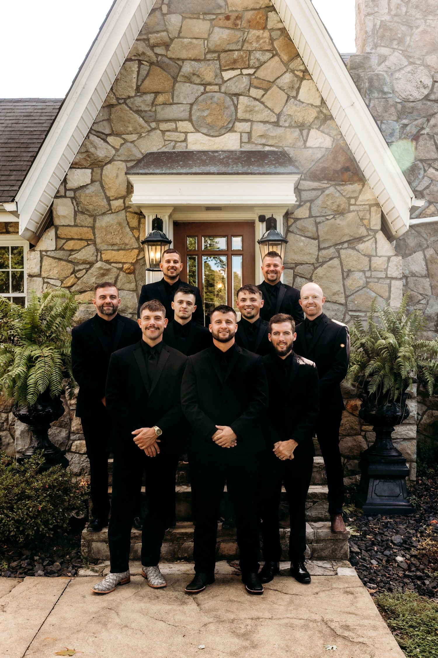 Groomsmen in matching black suits pose on stone steps outside an elegant stone building for a wedding photo.