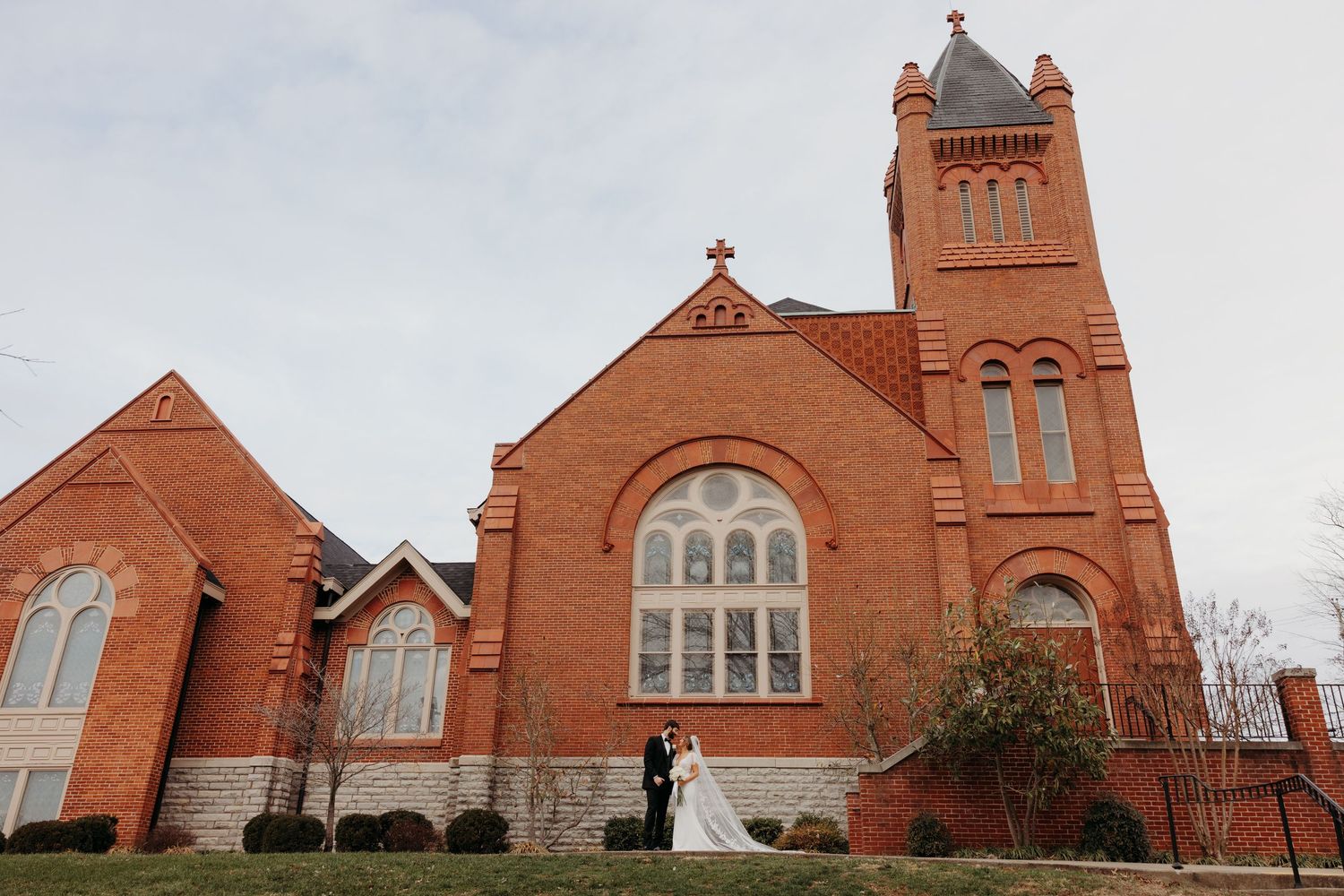 A historic red brick church with gothic architecture serves as backdrop for a wedding portrait.