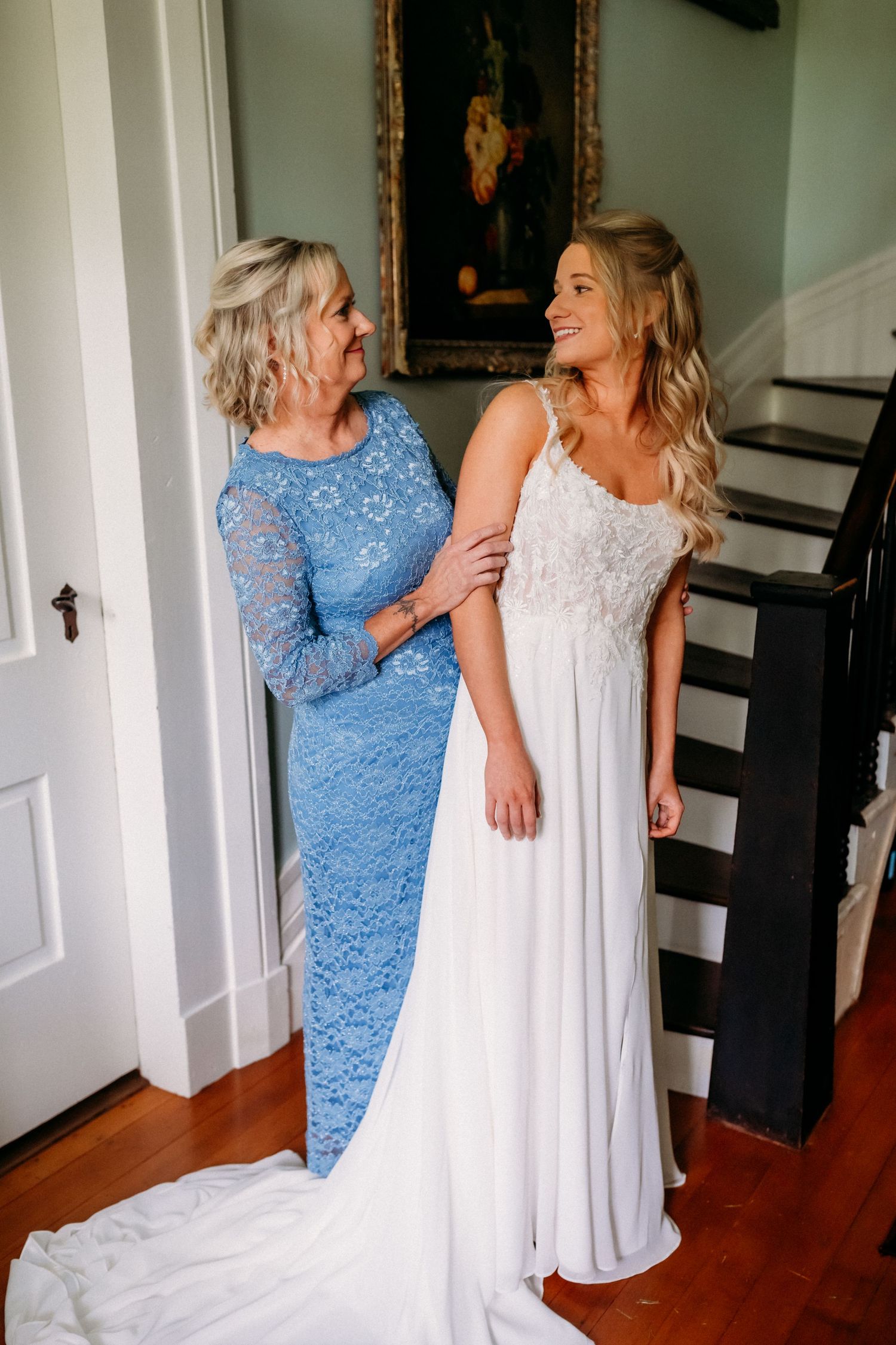 Bride in white lace wedding gown shares joyful moment with mother in blue lace dress before ceremony.