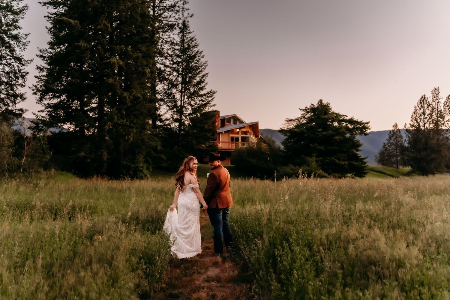 Bride and groom walk through tall grass meadow at golden hour near rustic lodge at western mountain wedding.