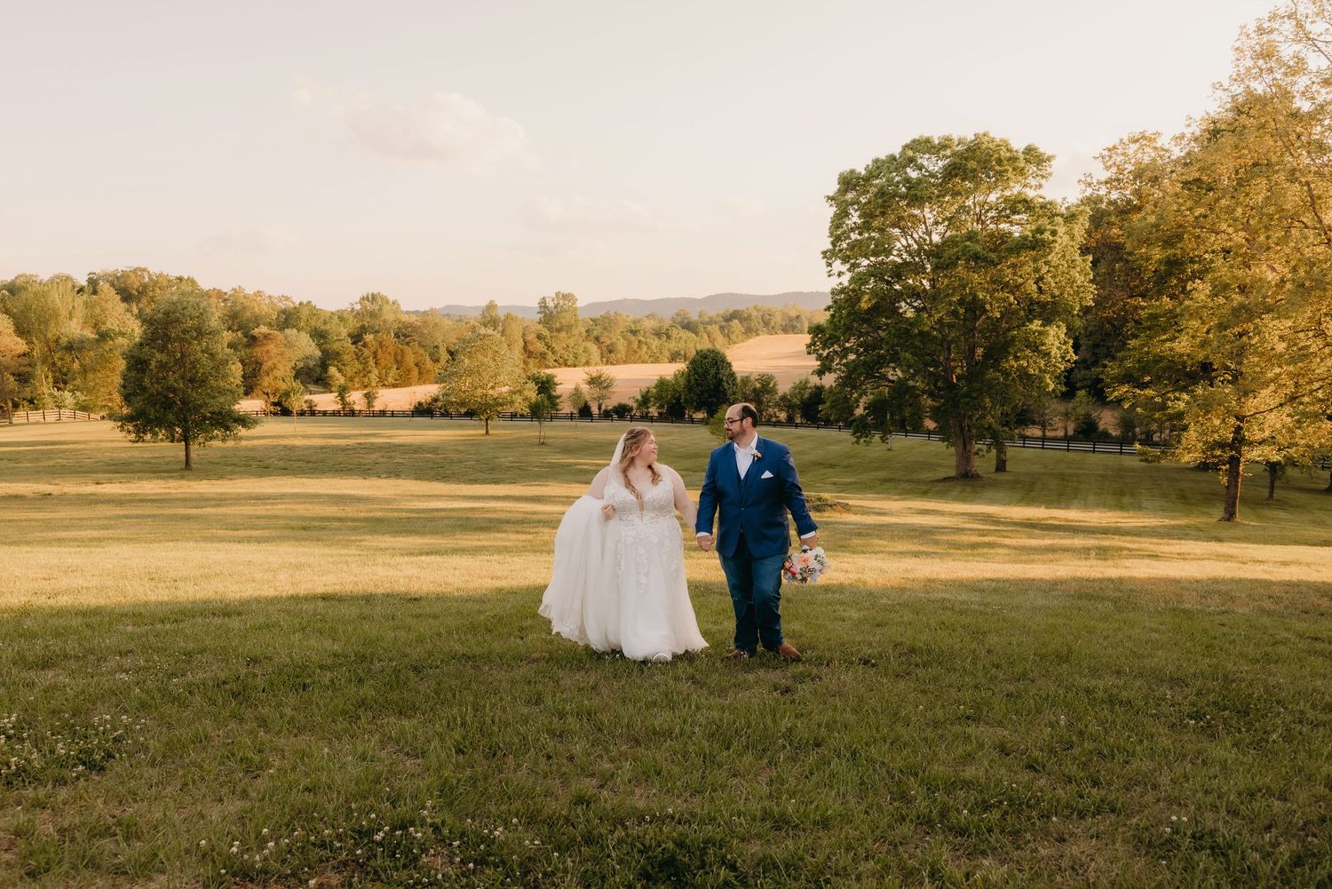Bride and groom walking hand in hand across a lush green field during golden hour at their outdoor wedding.