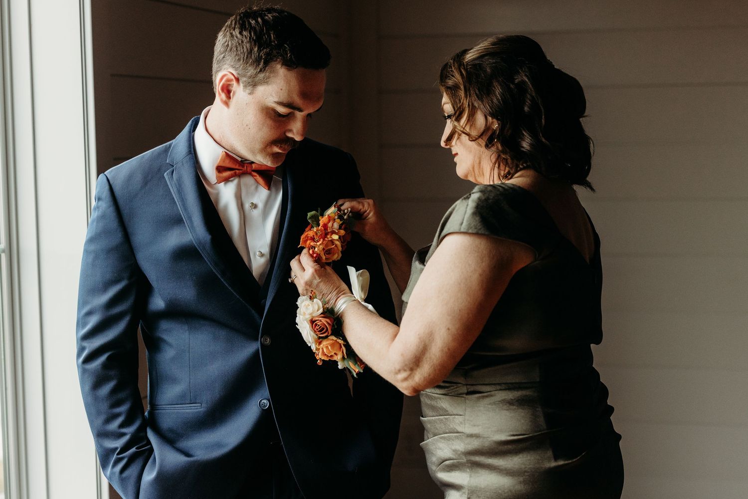 Woman pinning orange floral boutonniere on groom in navy suit and bow tie indoors.