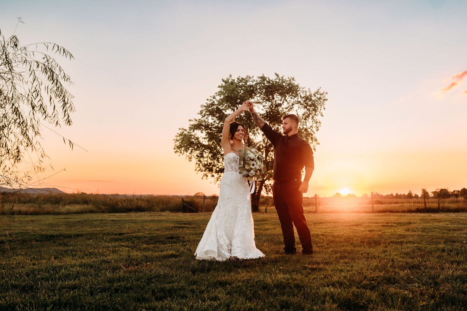 Bride and groom dancing at sunset in a green field with a tree and golden sky backdrop.