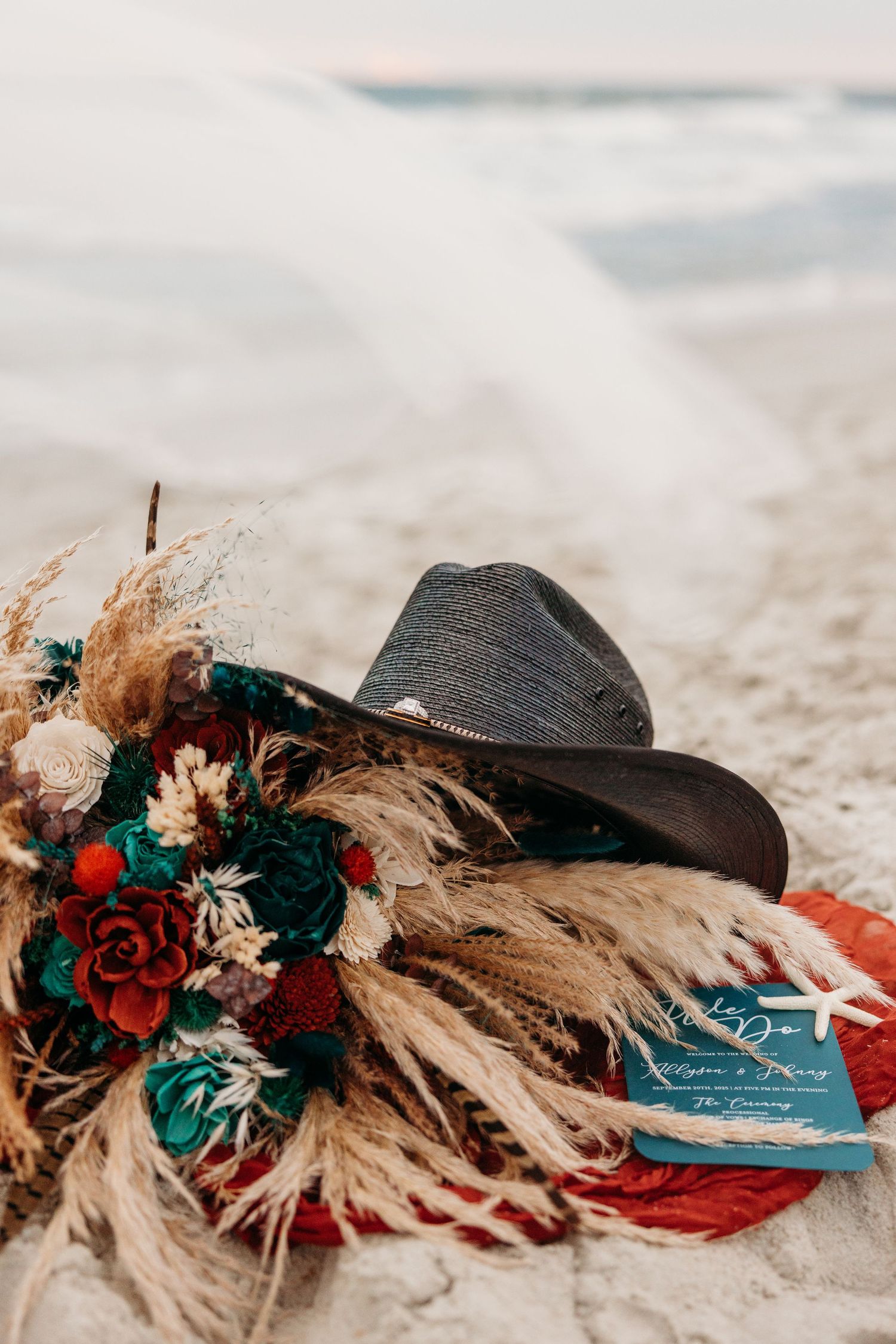 A black hat adorned with red roses and dried flowers lies on a sandy beach.