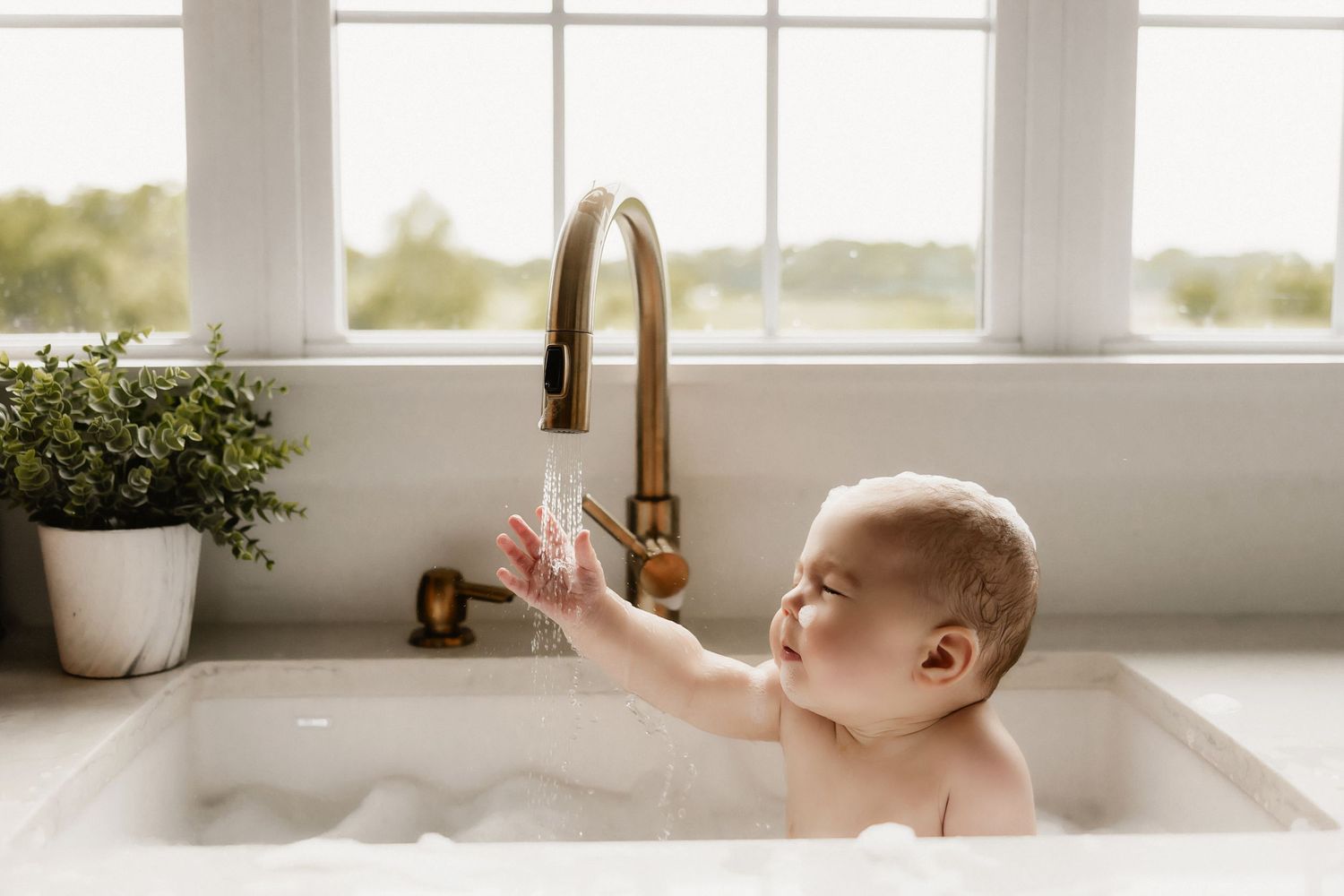 A toddler plays with water from a gold faucet in a bright white sink.