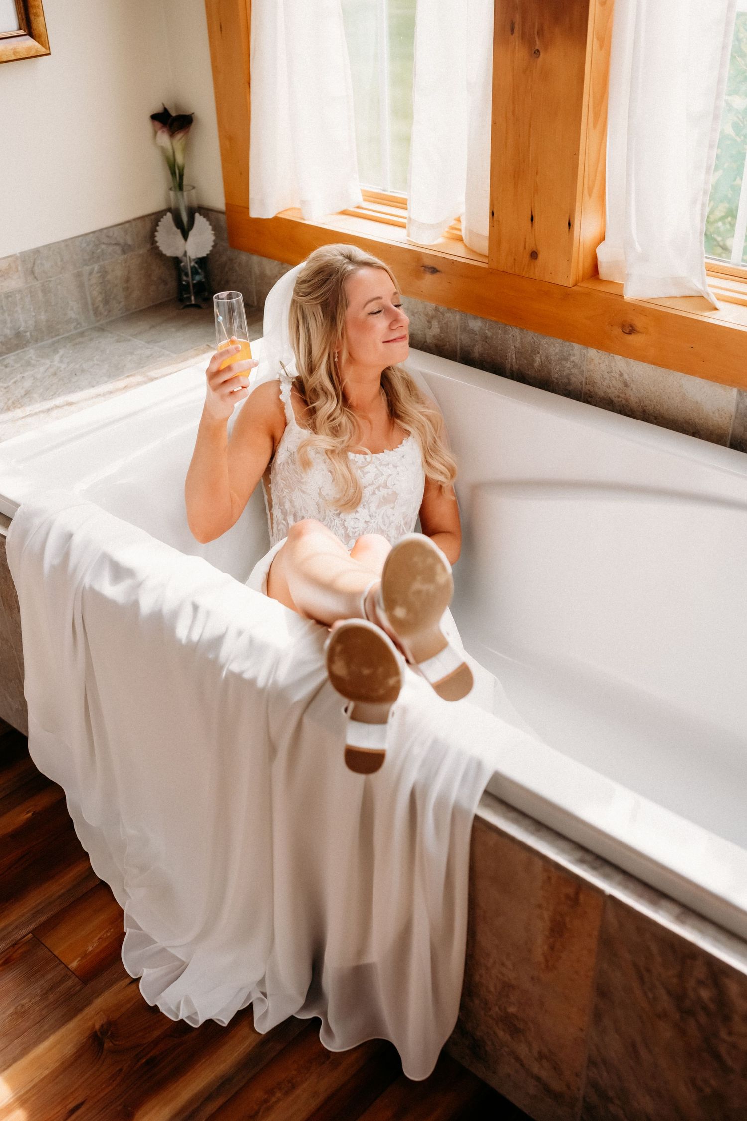 Smiling bride in wedding dress relaxing in bathtub holding champagne flute on wedding day.