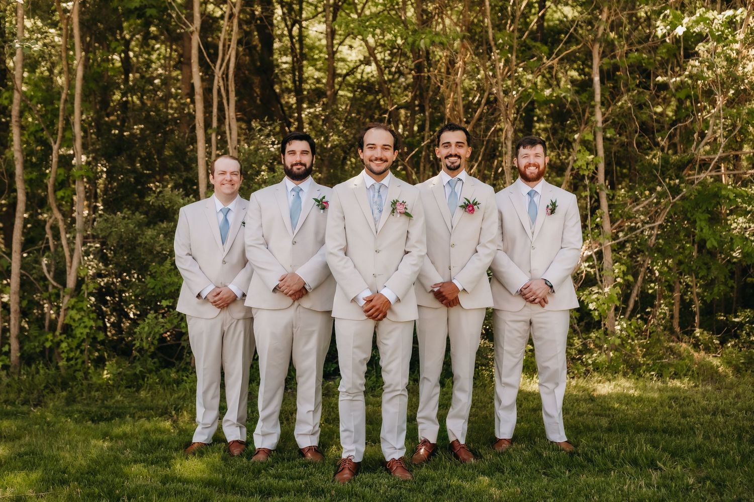 Groom and four groomsmen in matching tan suits pose outdoors in front of lush green trees.