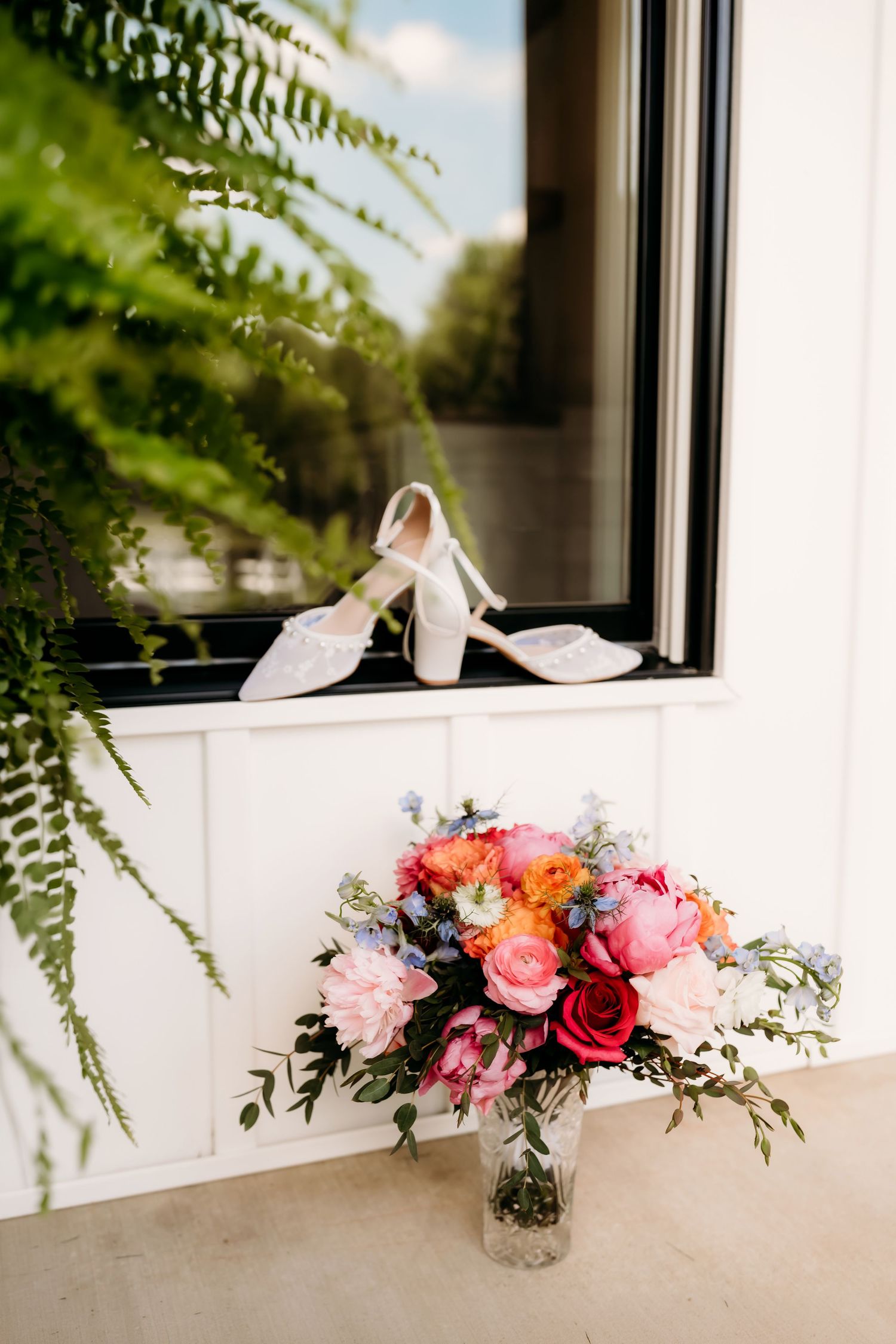 White bridal heels on windowsill beside colorful wedding bouquet with pink peonies, red roses, and blue flowers.