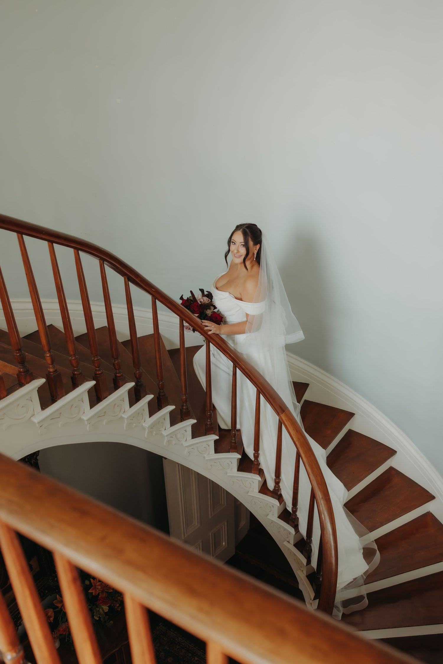 Bride in white off-shoulder gown and veil descending a curved wooden staircase with bouquet.