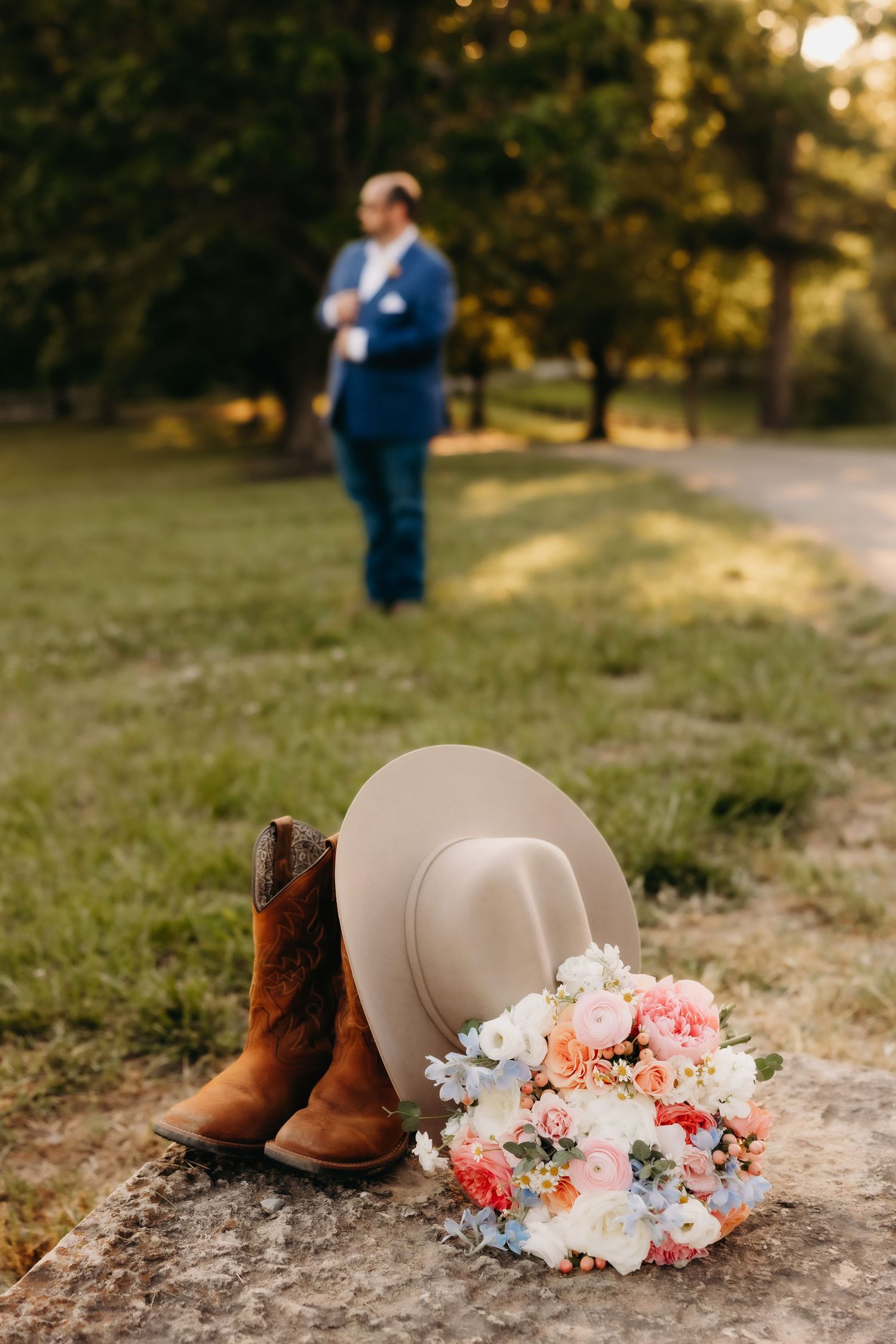 Cowboy hat, brown boots, and colorful bridal bouquet on stone with groom standing in background.