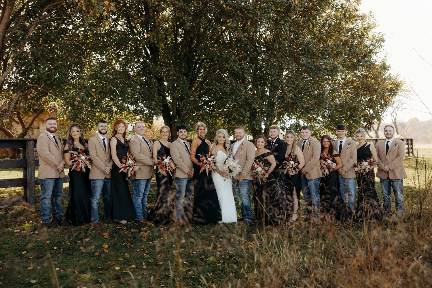 Wedding party in fall outdoor setting, bridesmaids in black floral gowns and groomsmen in tan blazers with jeans.