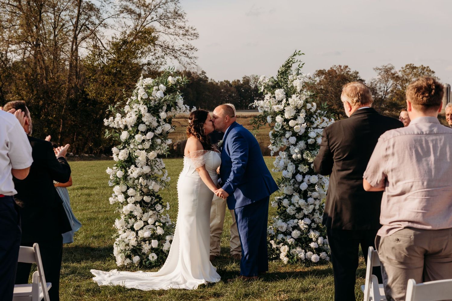 Bride and groom share first kiss at outdoor wedding ceremony with white floral arch on green lawn.