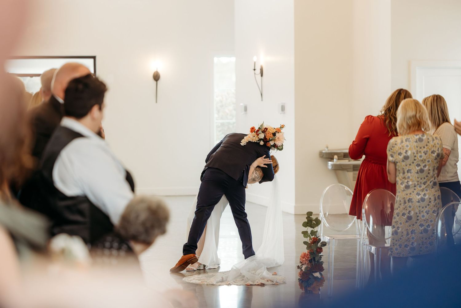 Groom bowing to bride during indoor wedding ceremony with guests seated and standing nearby.