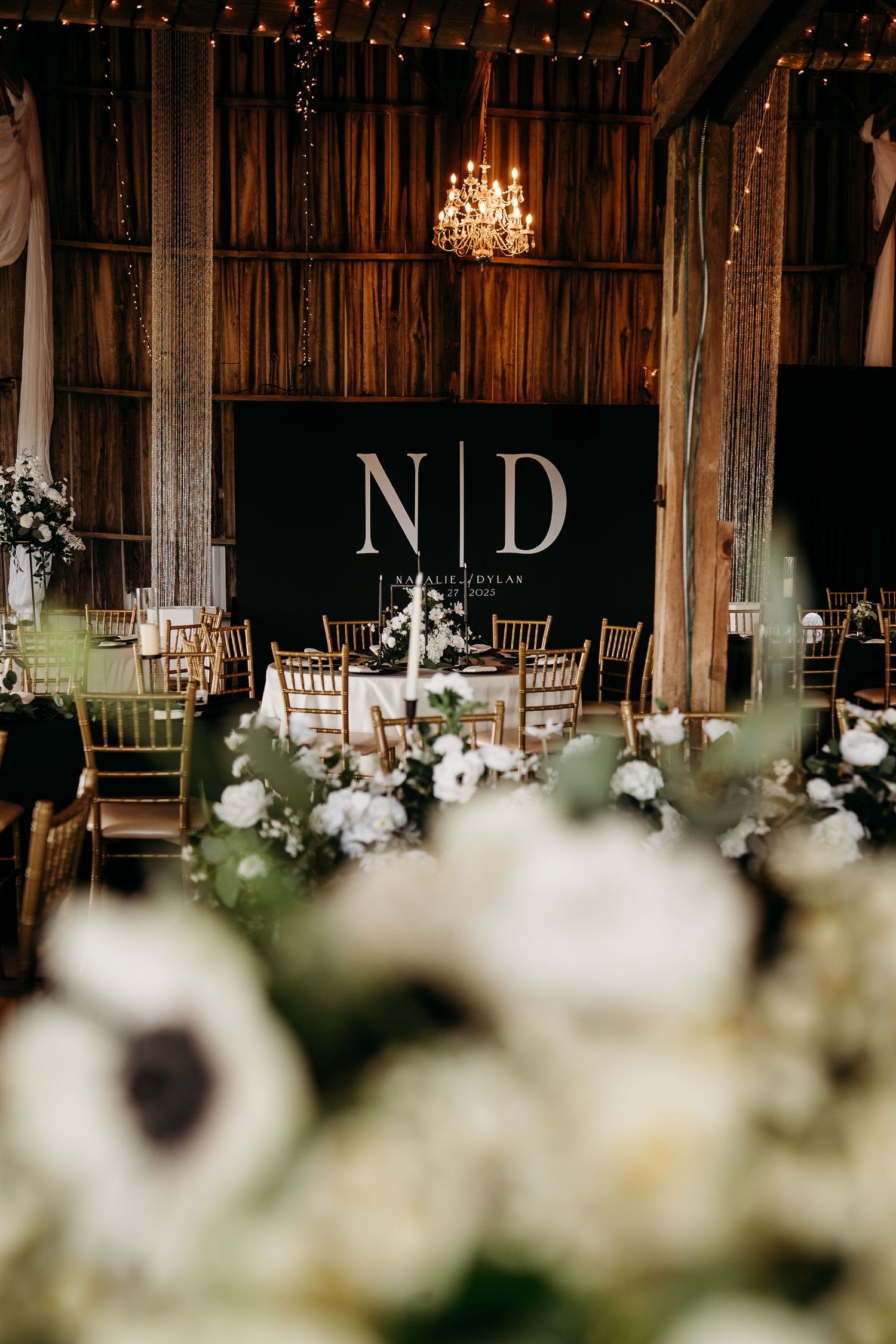 Elegant barn wedding reception with N&D monogram backdrop, chandelier, gold chairs, and white floral centerpieces.