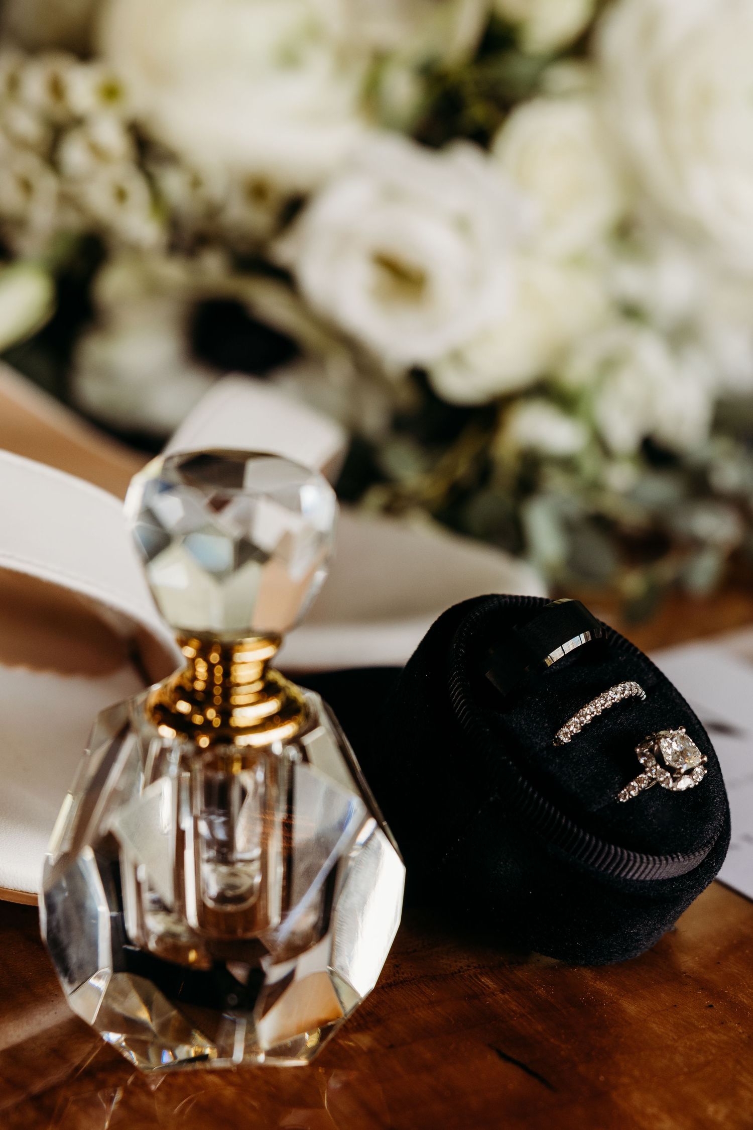 Crystal perfume bottle beside a black velvet ring box with diamond wedding rings, surrounded by white roses.