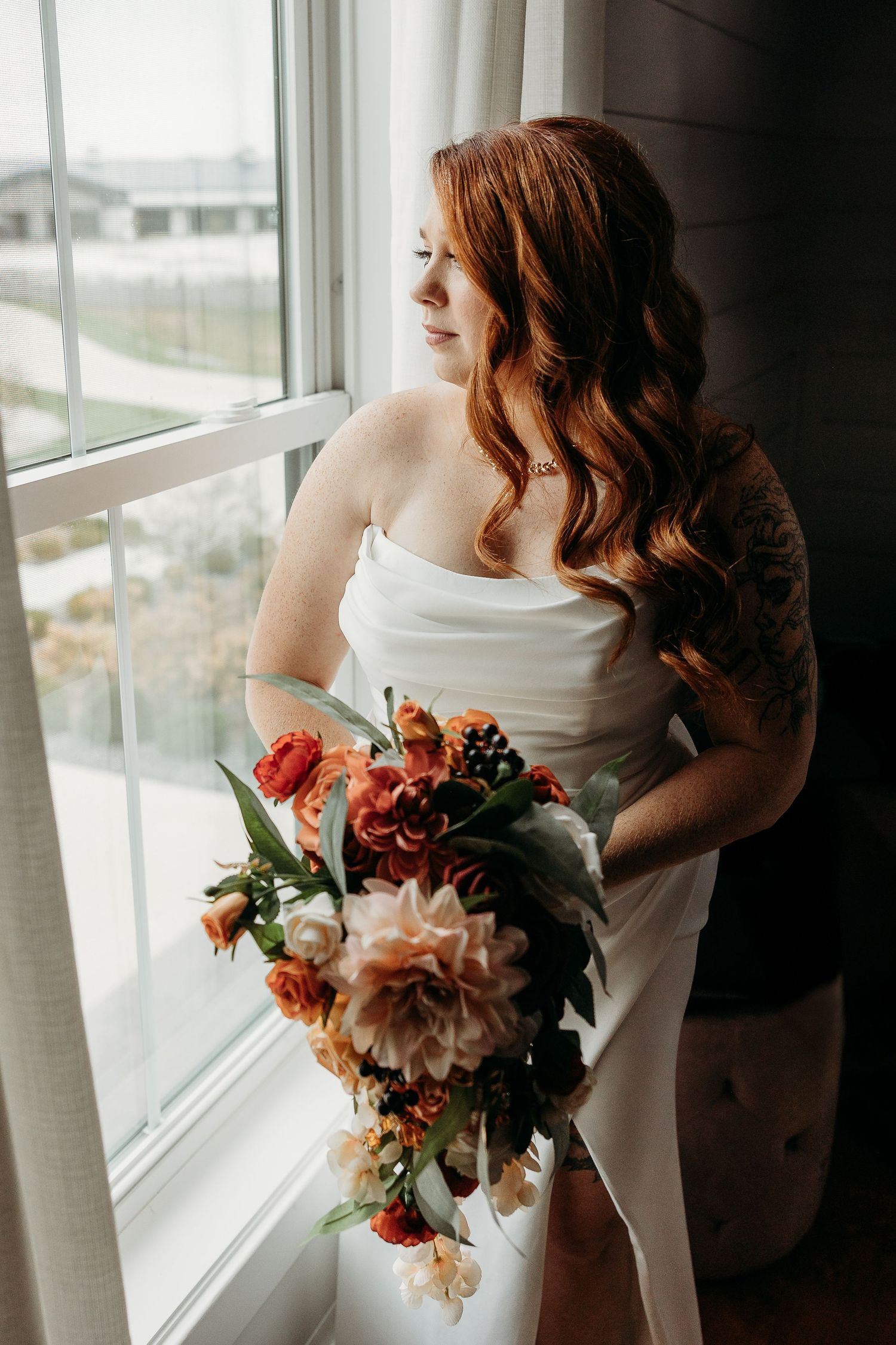 Bride in white gown holds colorful bouquet of fall flowers by window on her wedding day.