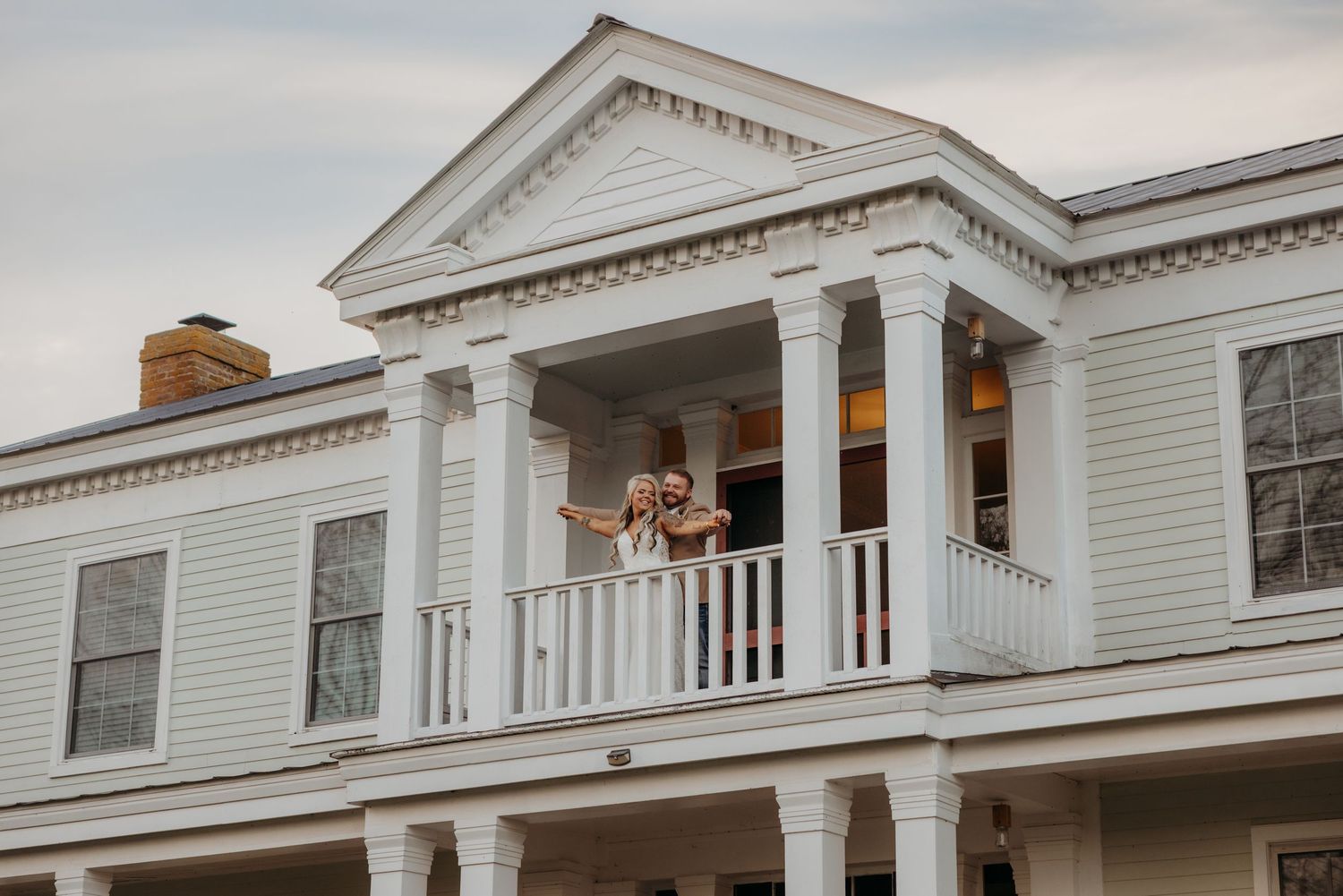 Couple waving from white colonial mansion balcony at outdoor wedding venue at dusk.