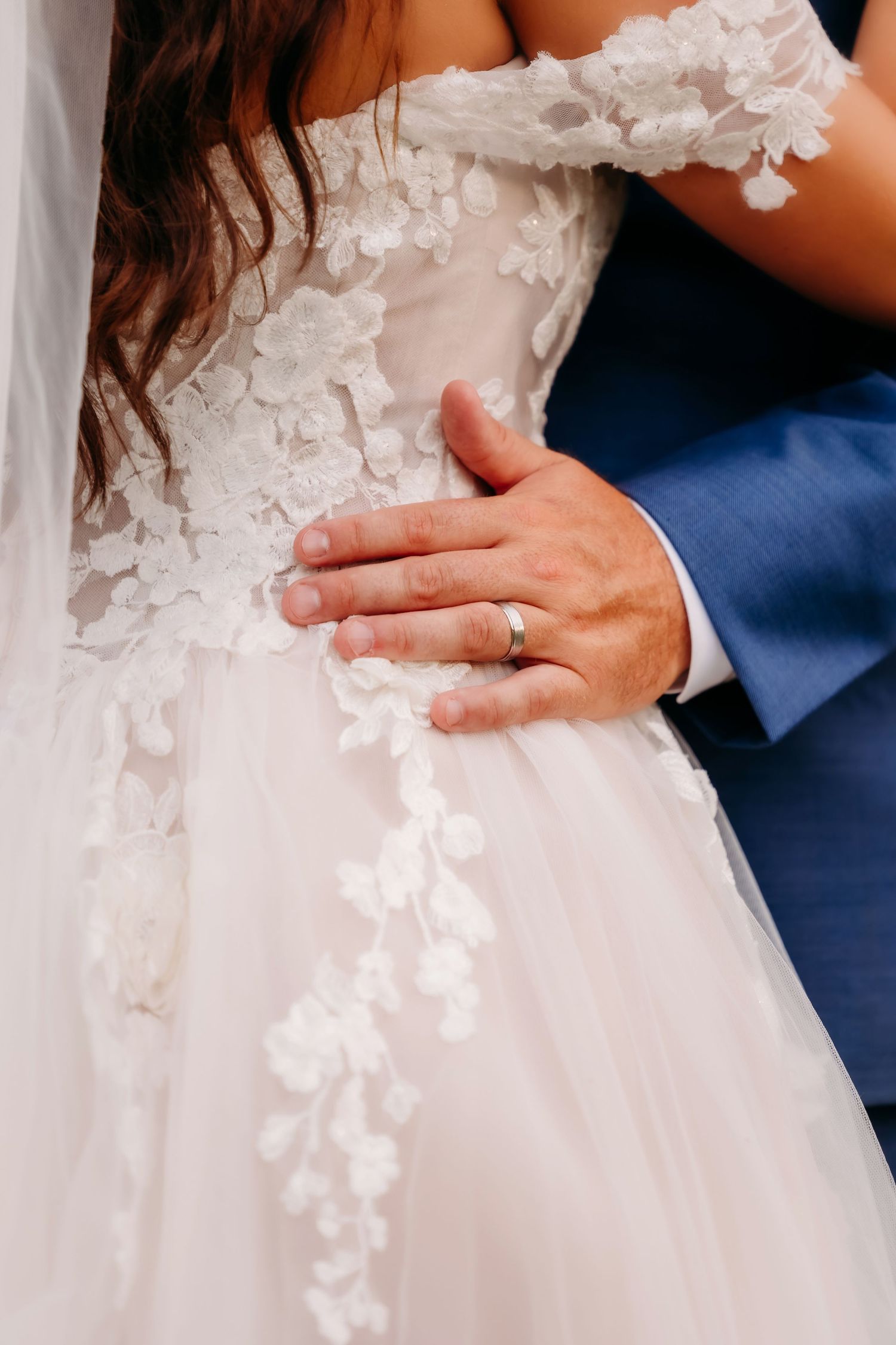 Groom's hand with gold wedding band resting on bride's intricate floral lace wedding dress.