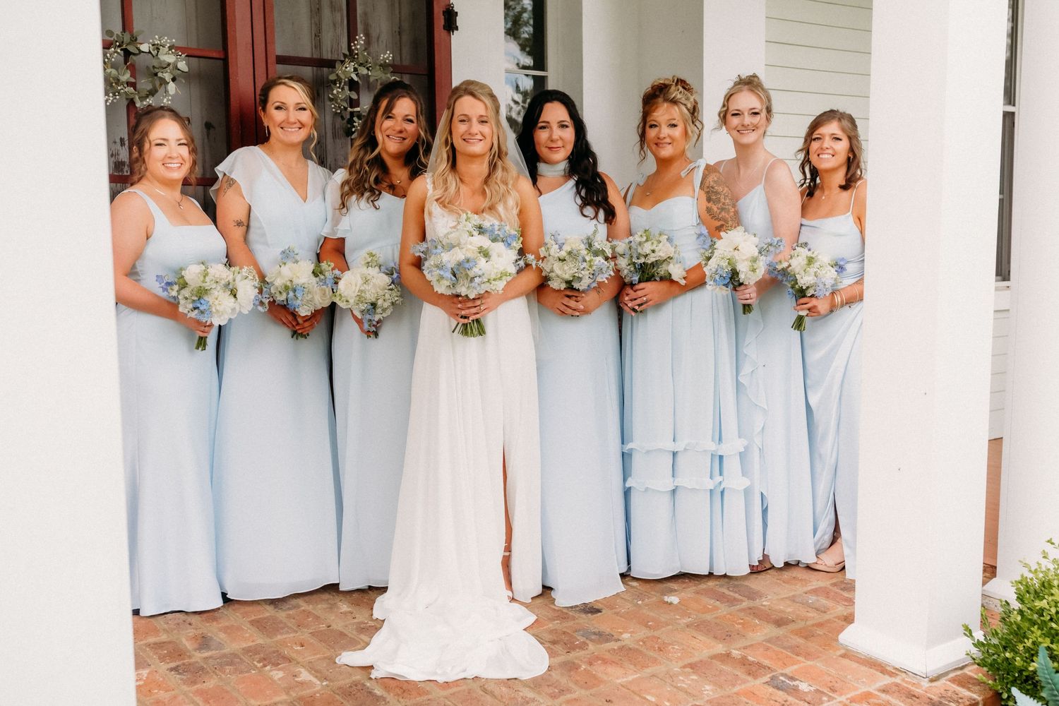 Bride in white gown poses with six bridesmaids in light blue dresses holding white floral bouquets on a brick porch.
