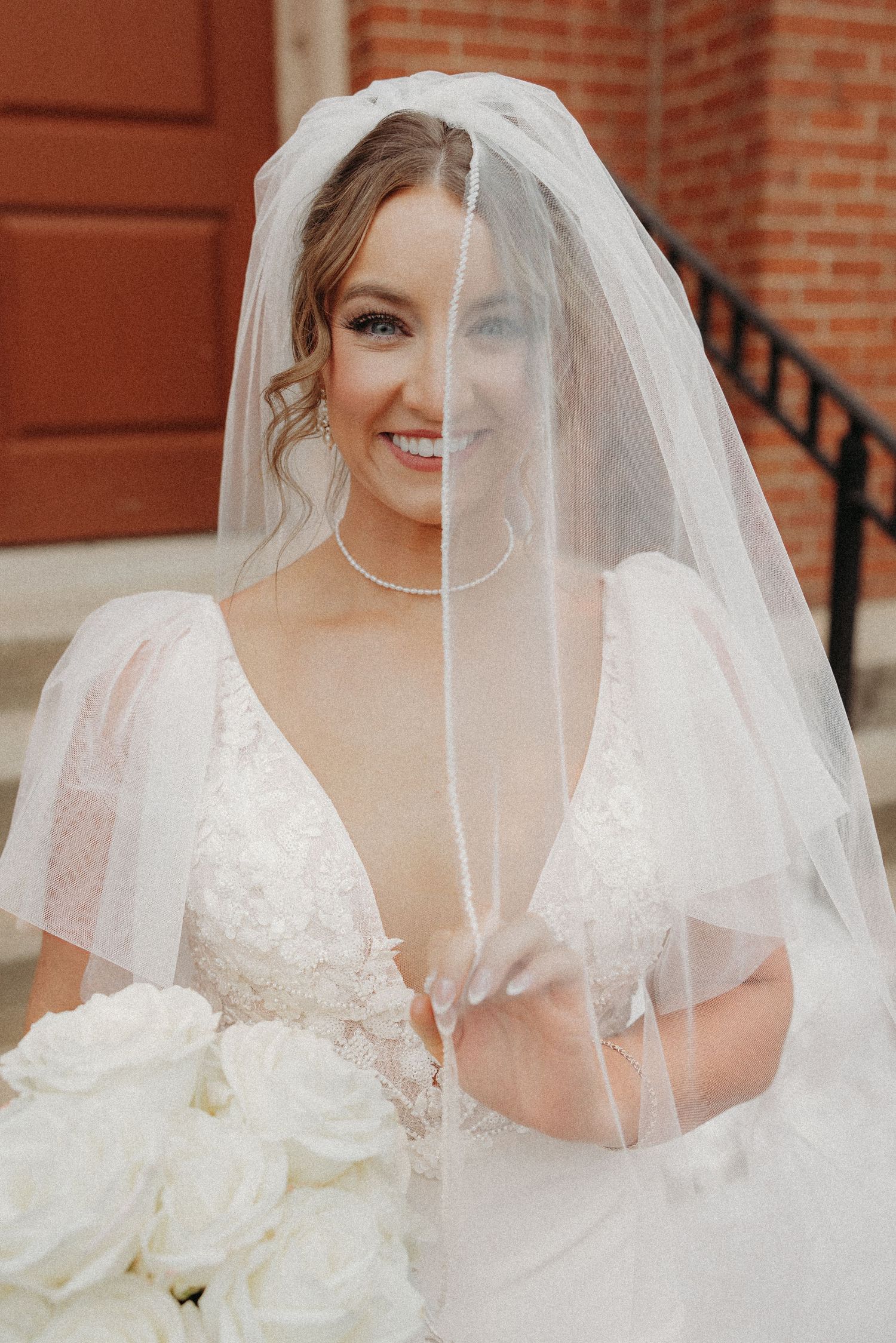 Smiling bride in lace V-neck gown holds white rose bouquet with veil draped over face outside brick church.