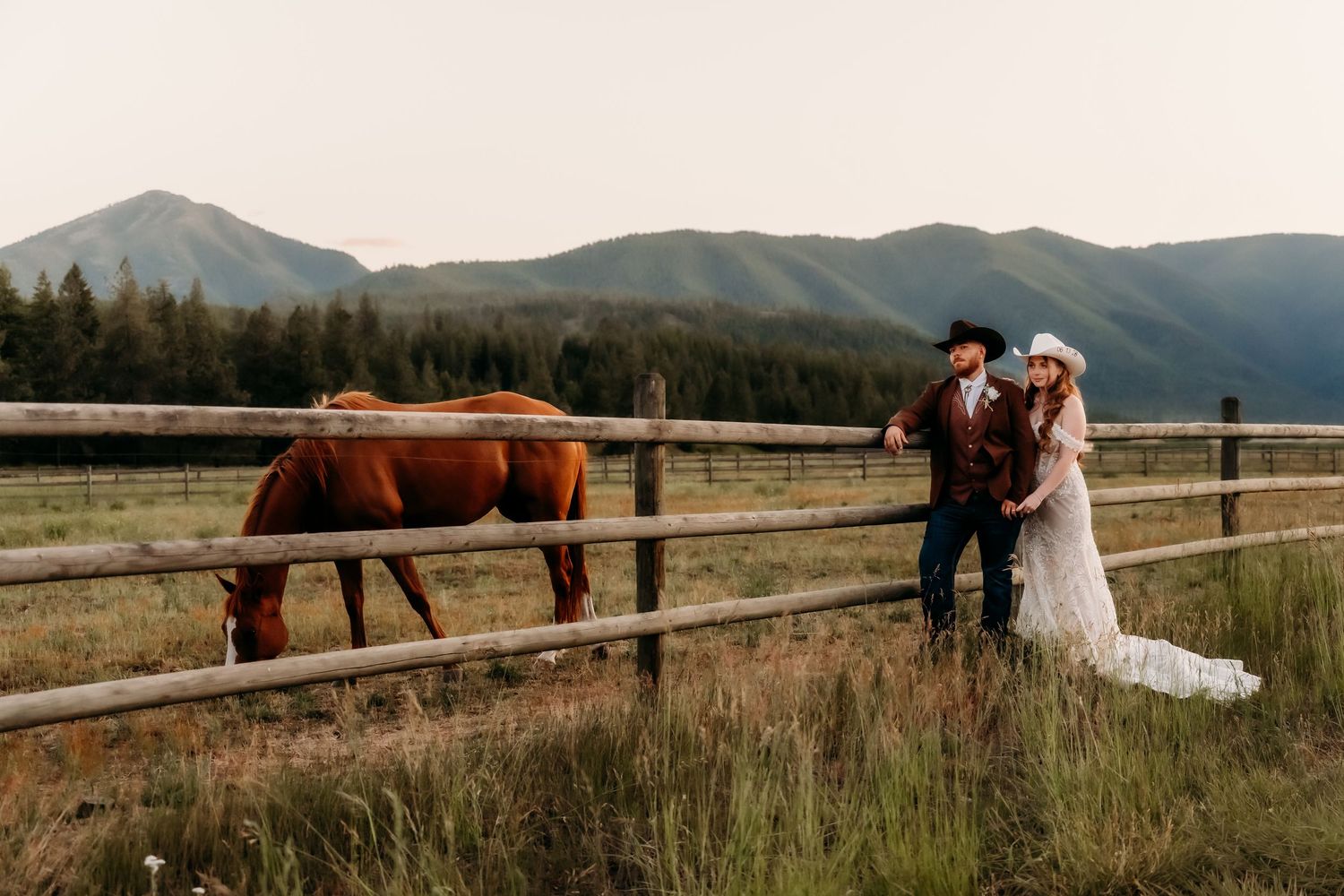 Bride and groom in western attire pose by a wooden fence with a chestnut horse and mountain backdrop.