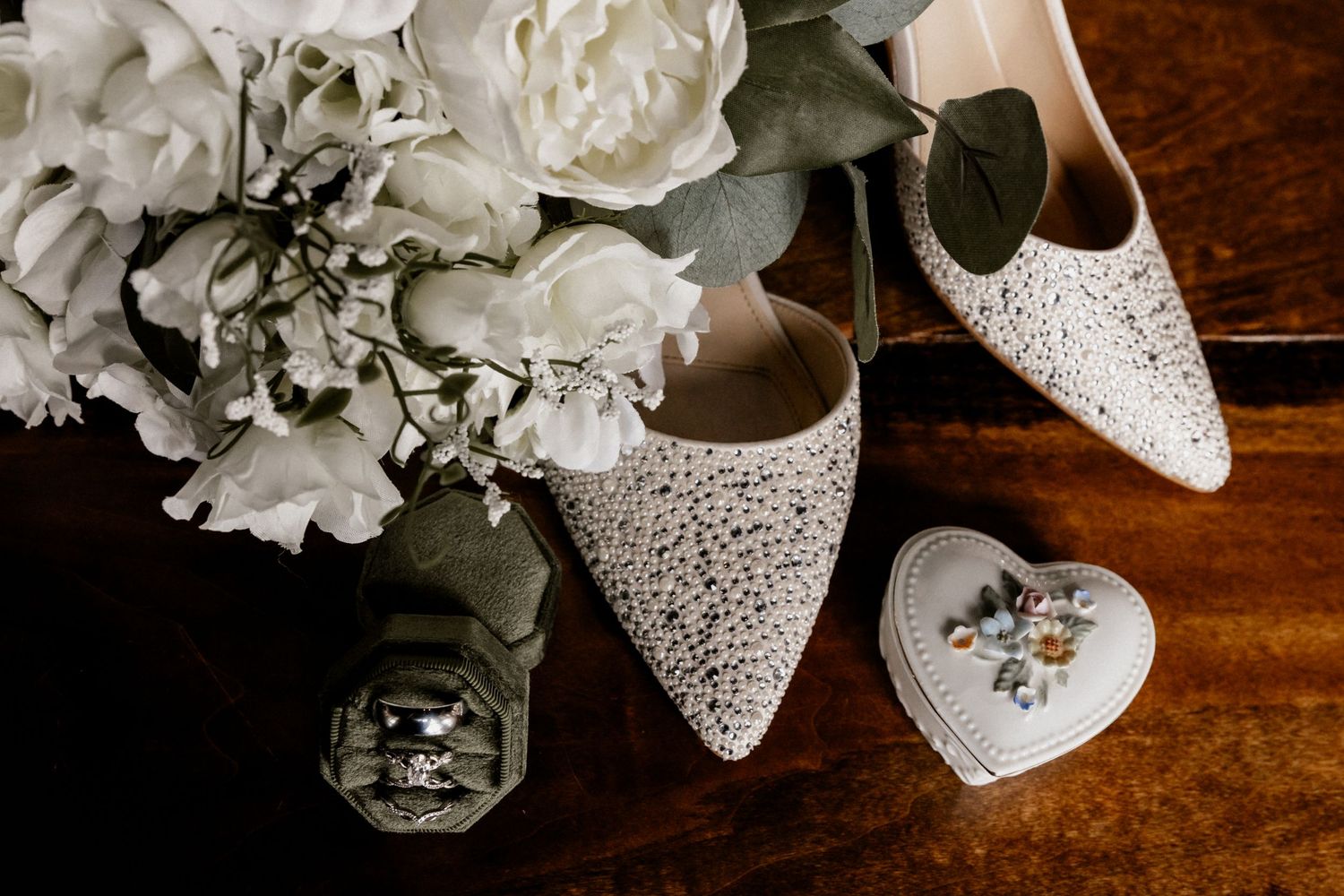 Wedding accessories including white shoes, flowers, and jewelry arranged on dark wooden surface.