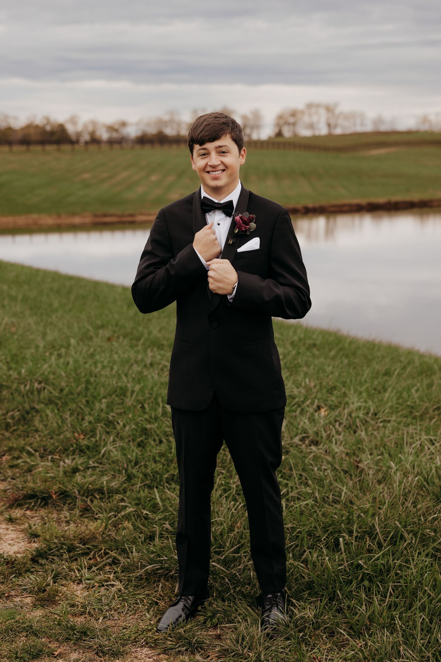 Groom in black tuxedo poses playfully on grassy field near pond at Simmstown Manor wedding venue in Springfield, KY.