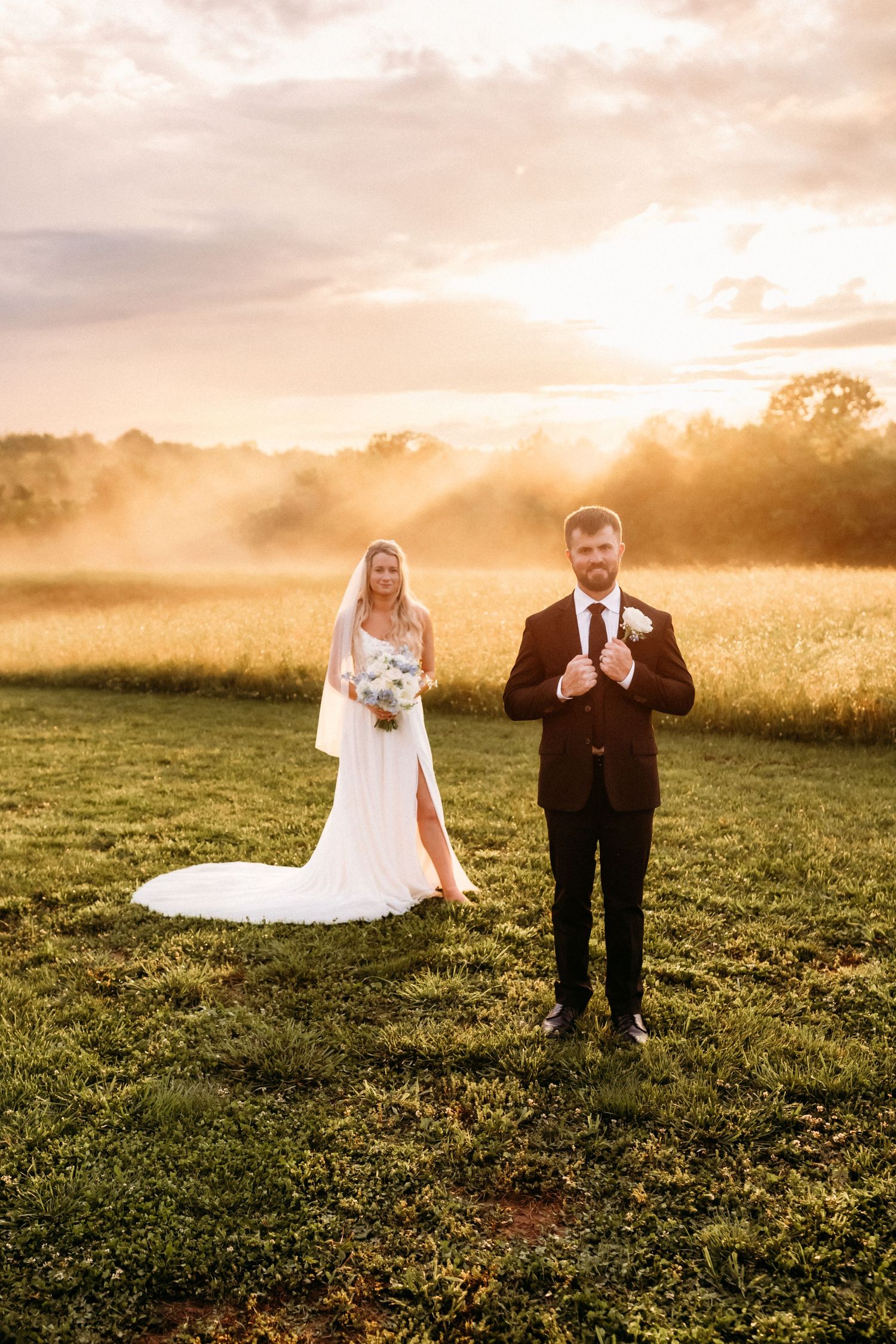 Bride in white gown and groom in black suit pose at golden sunset in misty green field.