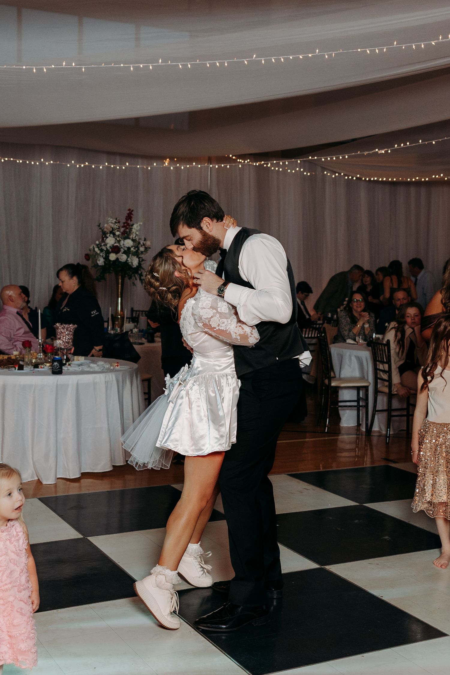 Bride and groom share a romantic first dance kiss on a black and white checkered dance floor at their wedding reception.