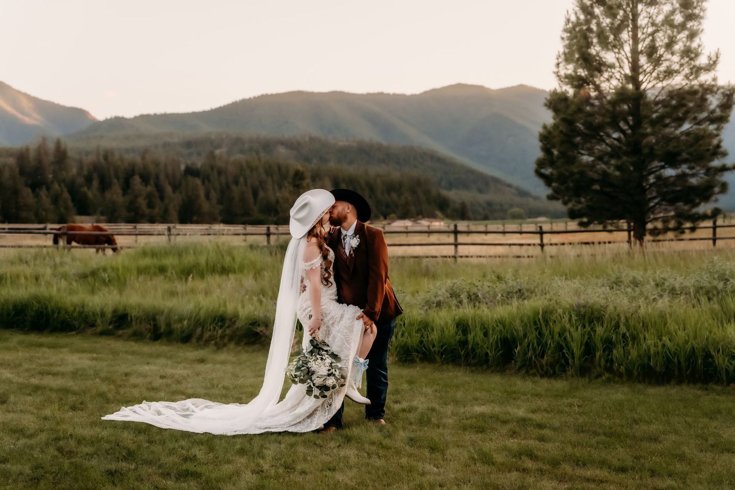 Bride and groom share romantic dip kiss in mountain meadow at golden hour with horses and pine trees.