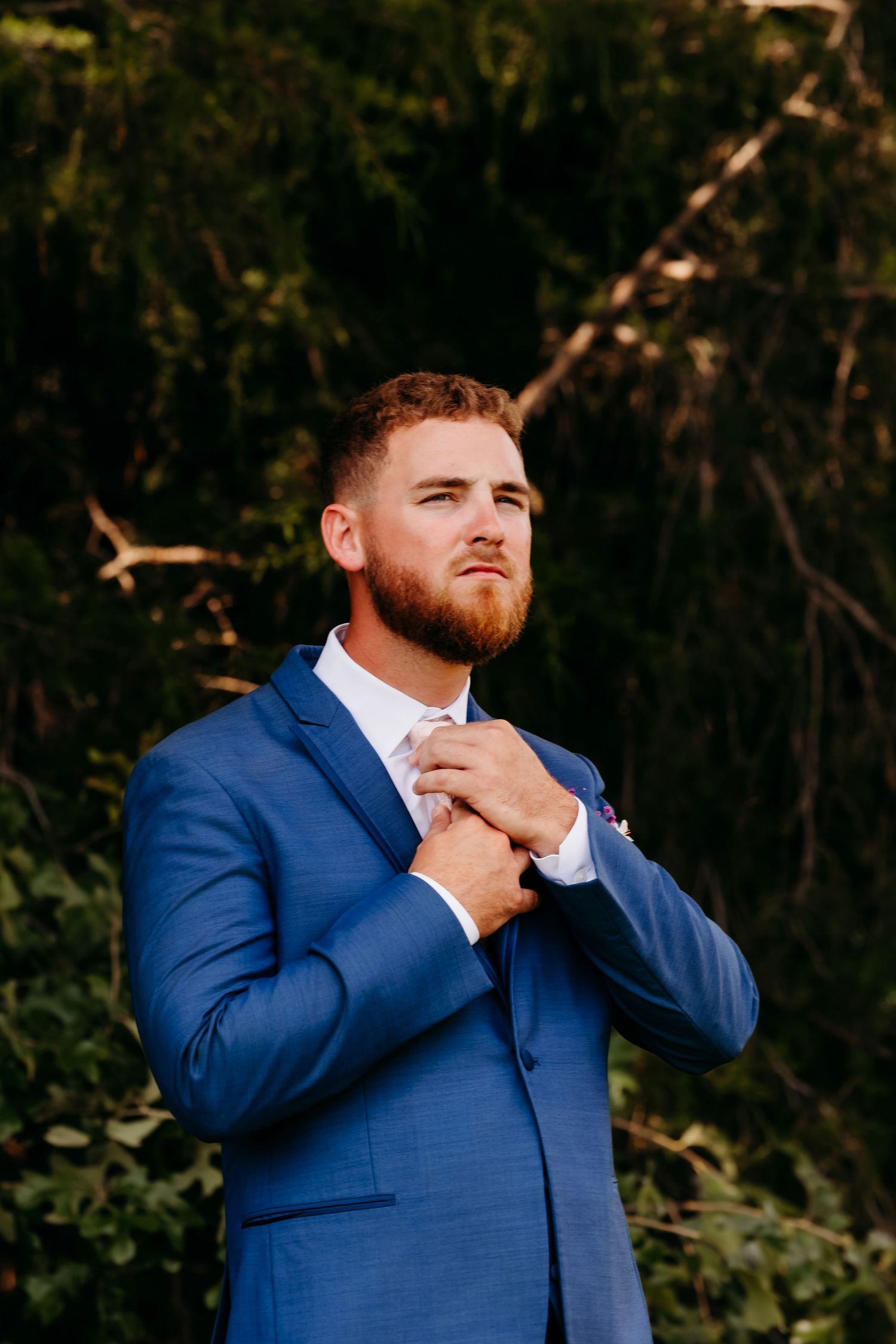 Handsome groom in blue suit adjusting his tie outdoors with lush green trees in the background.